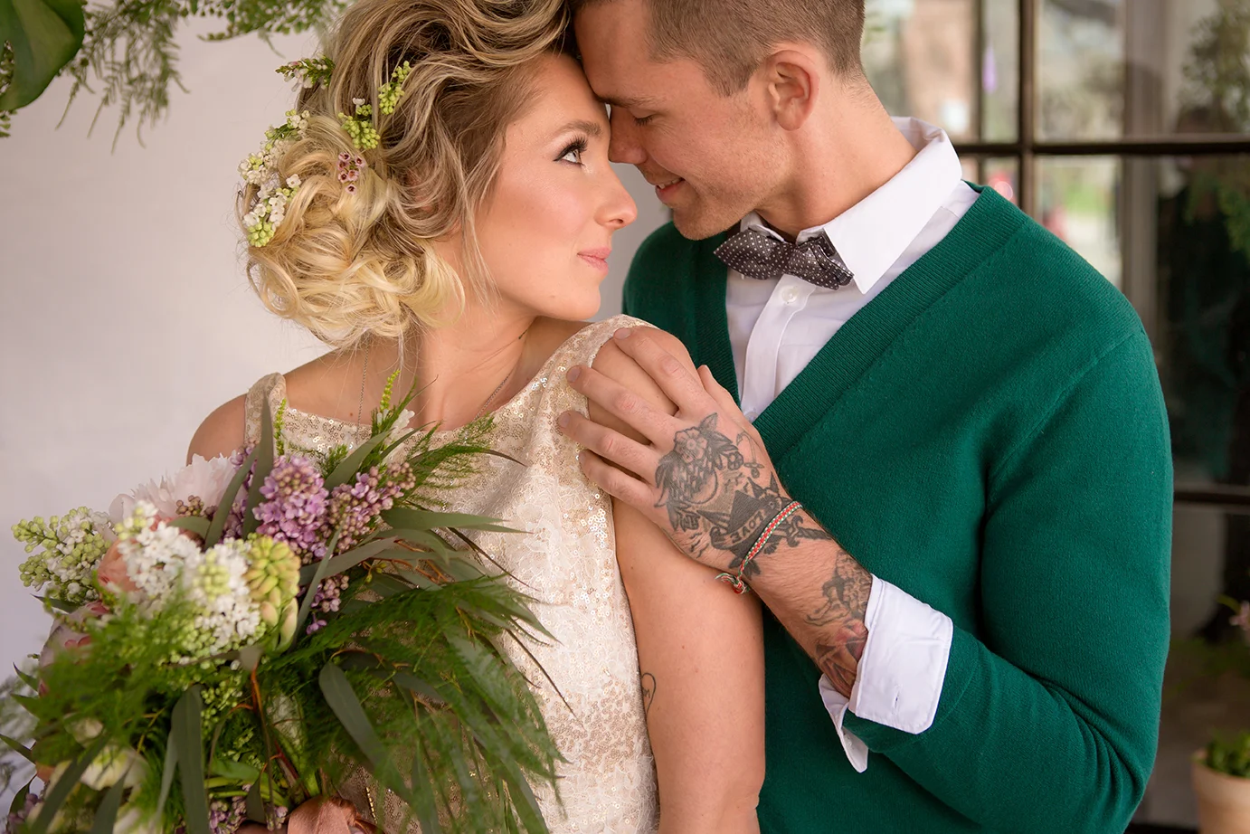 A couple, a woman in a floral dress holding a bouquet and a man in a green sweater and bow tie, touching foreheads with eyes closed, smiling, in an indoor setting with window behind.
