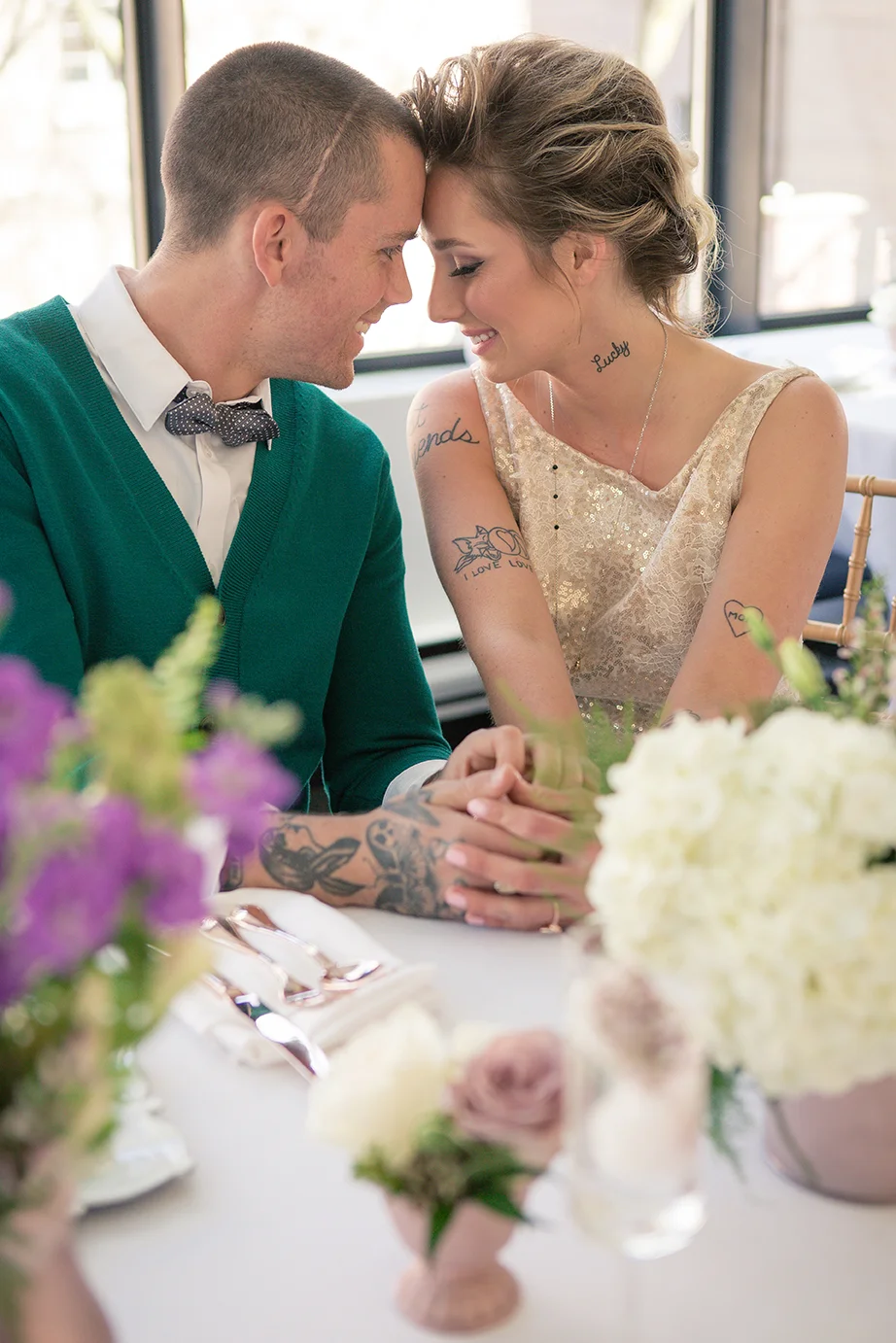 A happy couple sitting closely at a table, holding hands and touching foreheads, during a celebration or special event, with flowers on the table.