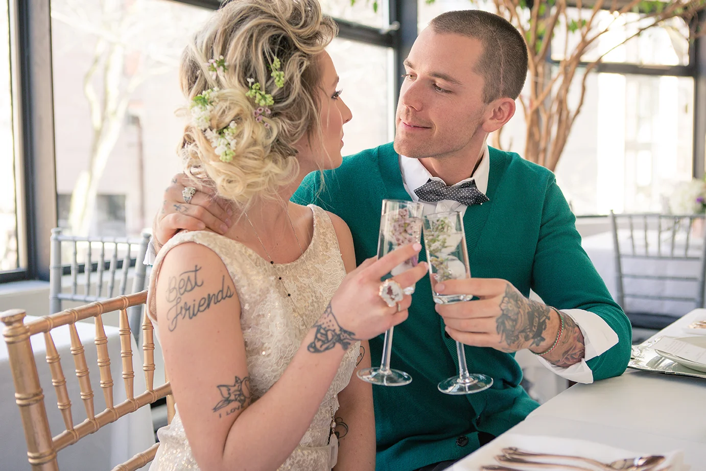 A bride and groom sitting close at a wedding reception, holding champagne glasses and gazing at each other in a bright, decorated venue with large windows.