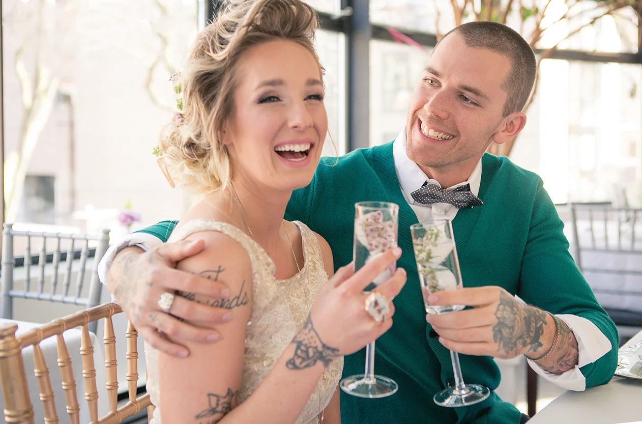 A smiling woman with tattoos on her arm sitting next to a man with tattoos, holding glasses of water with floral decor inside, during a celebration or special occasion.