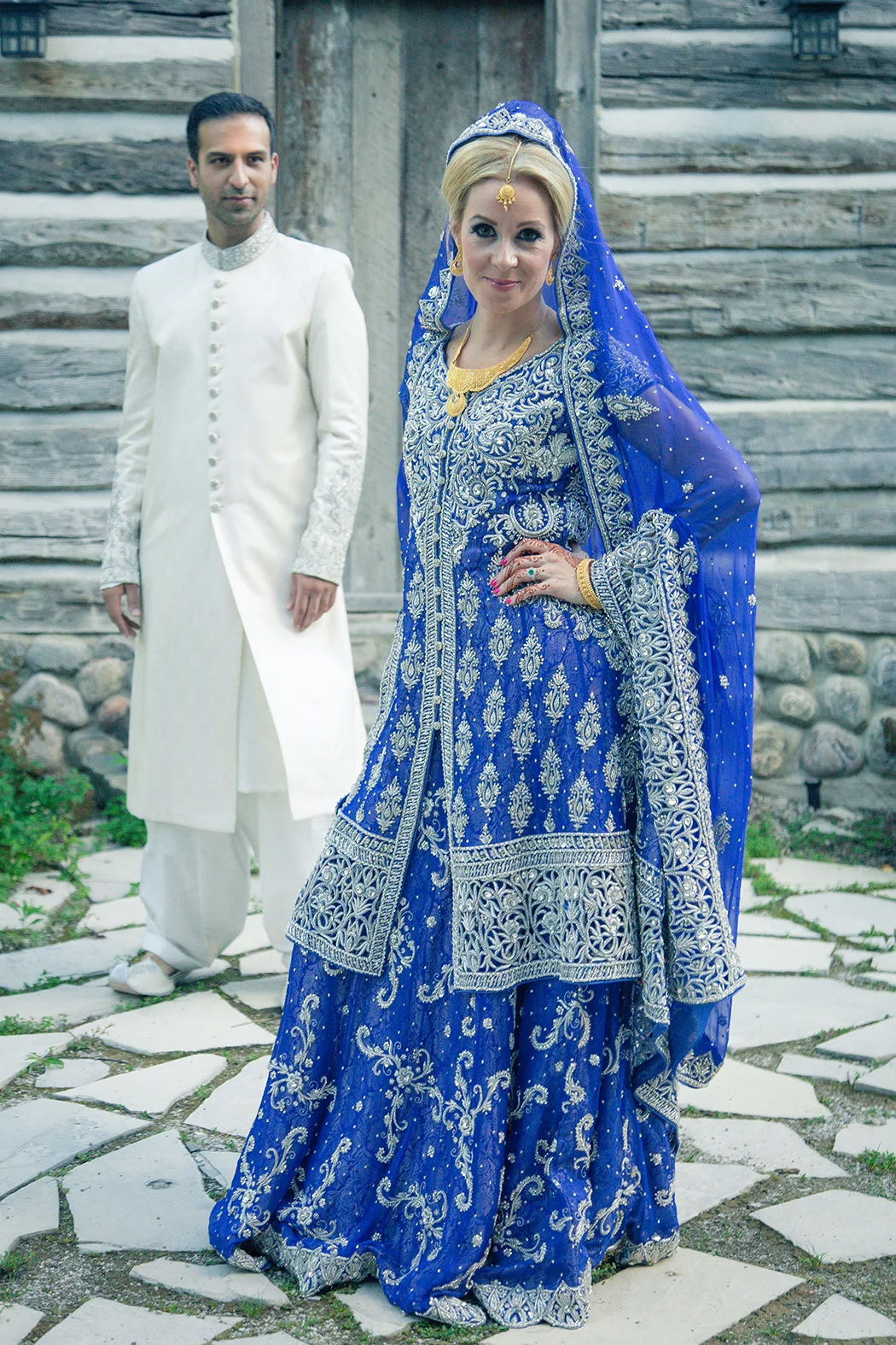 A woman wearing an elaborate blue traditional South Asian dress with silver embroidery, gold jewelry, and a sheer veil, standing outdoors on a stone pathway. A man in a white traditional outfit stands in the background.