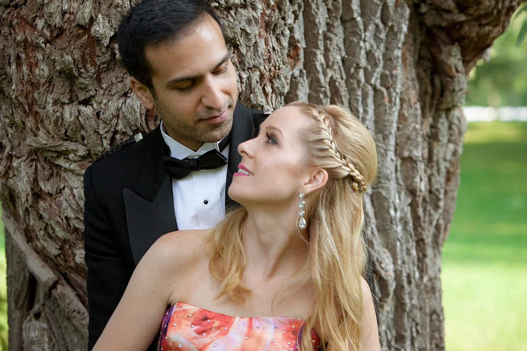 A man in a tuxedo and a woman in a colorful strapless dress pose close to each other outdoors with a large tree in the background.