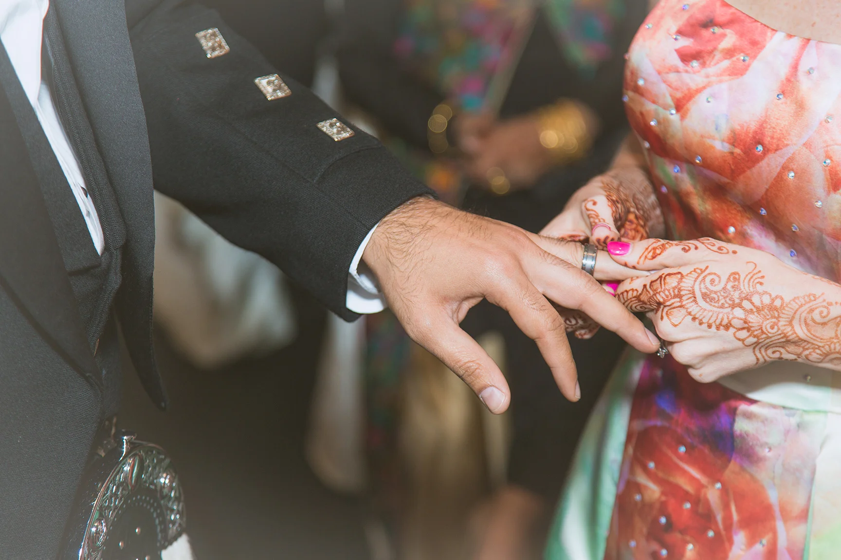 A close-up of a wedding ceremony showing a couple exchanging rings, with the groom's hand over the bride's hand. The groom is wearing a black suit with decorative buttons on the sleeve, and the bride's hands are adorned with henna designs. The bride 