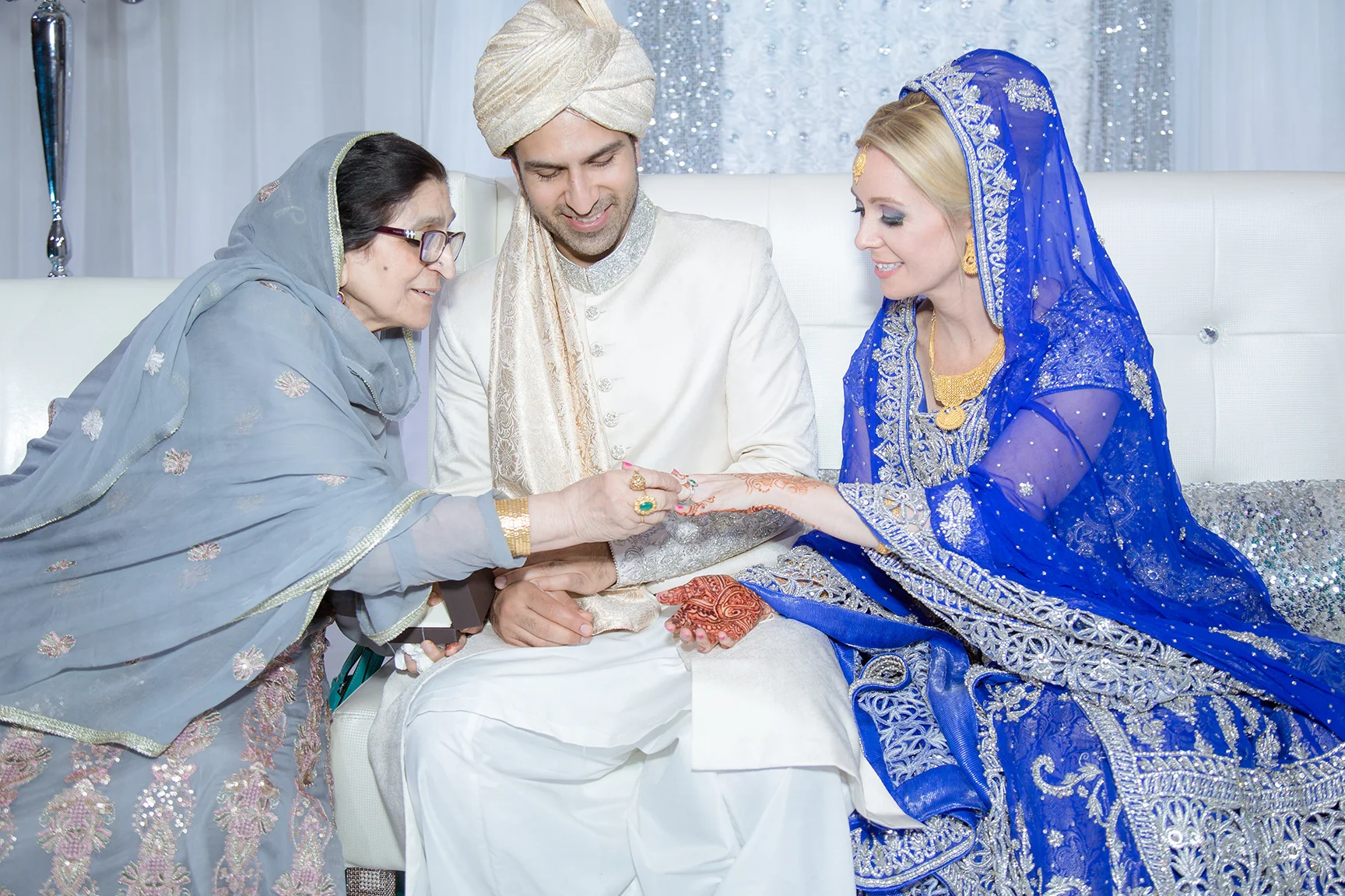 Indian wedding ceremony with bride and groom exchanging flower garlands, woman in blue traditional attire, man in cream sherwani, woman in grey sari observing.