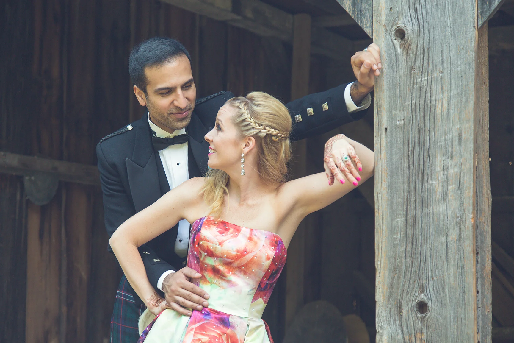 A happy couple, the man in a black tuxedo and bow tie, and the woman in a colorful strapless dress, enjoy a romantic moment outdoors, leaning against a wooden post.
