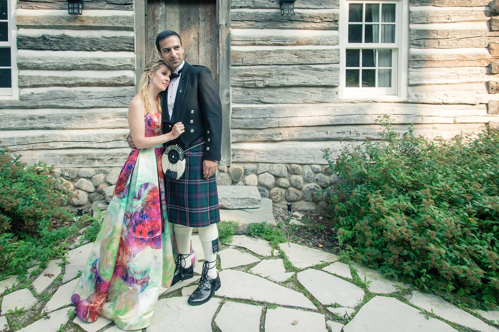 A couple dressed in formal and traditional Scottish attire standing outside a rustic wooden house with stone foundation, green shrubs, and stone pathway.