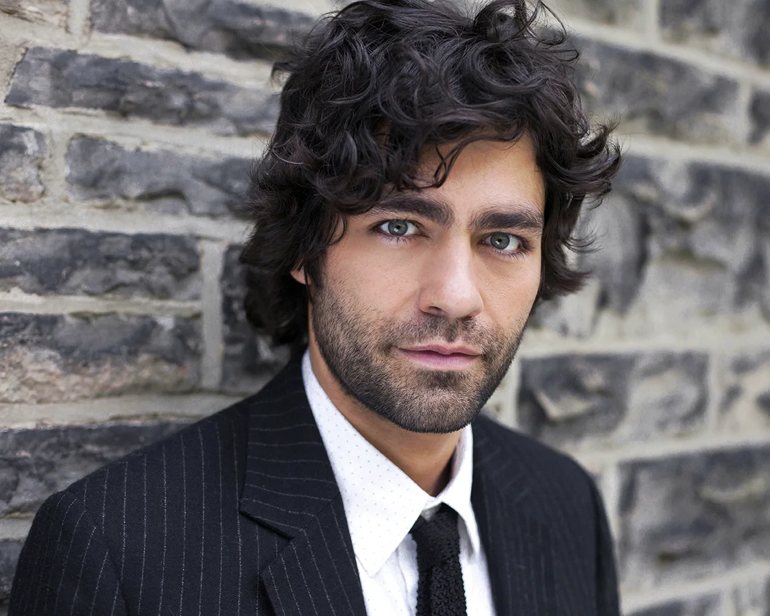 A man with curly dark hair and blue eyes, wearing a black pinstripe suit, white dotted shirt, and black tie, standing against a brick wall.