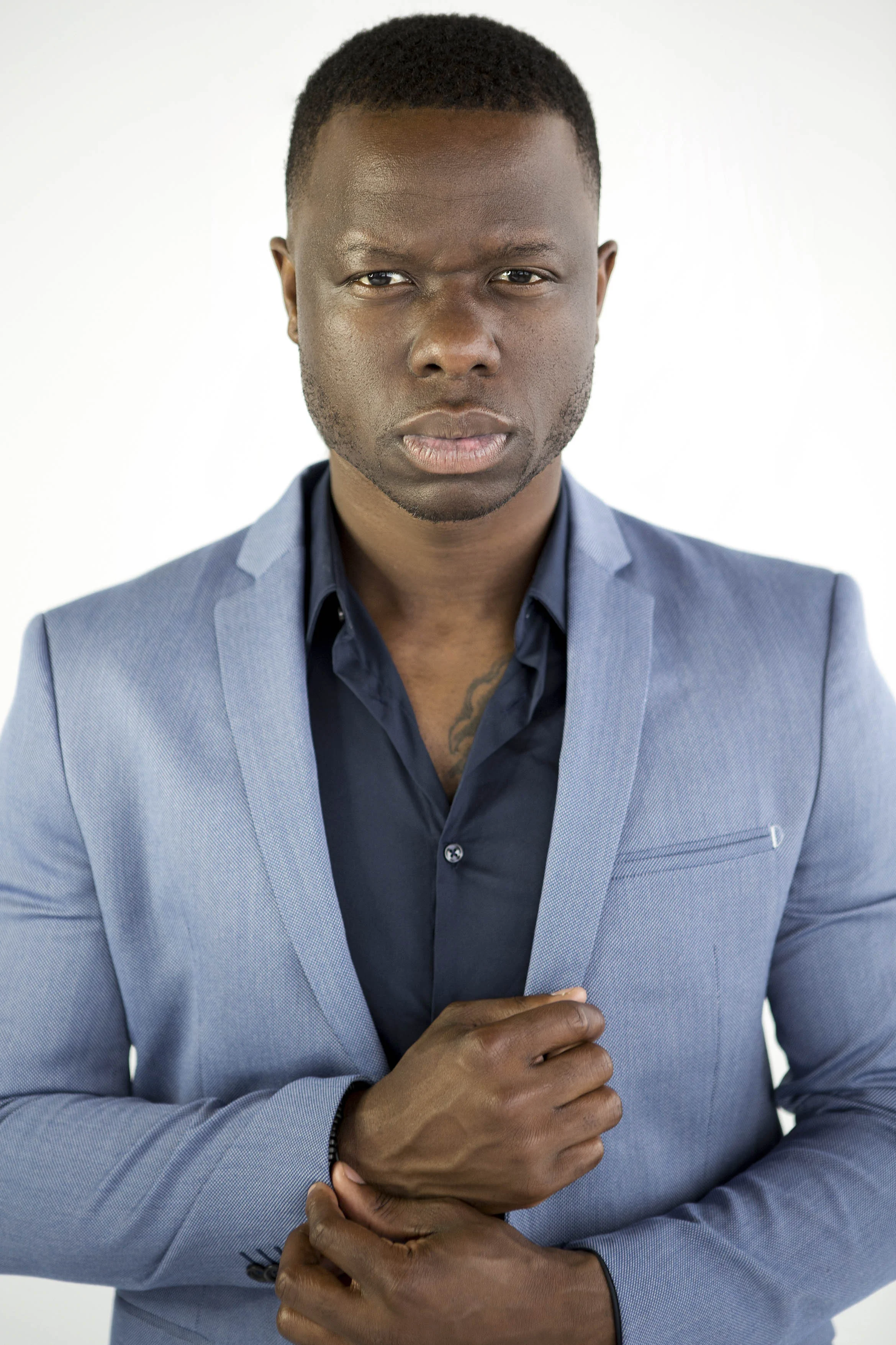 Portrait of a serious young Black man with short hair wearing a light blue blazer and a dark blue shirt, standing against a plain white background.