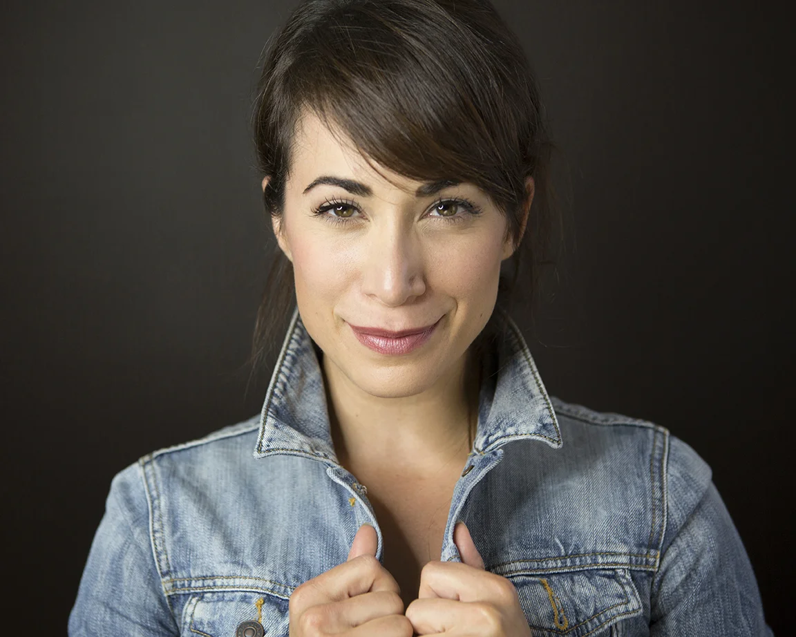 A woman with short brown hair, wearing a denim jacket, smiling gently against a dark background.