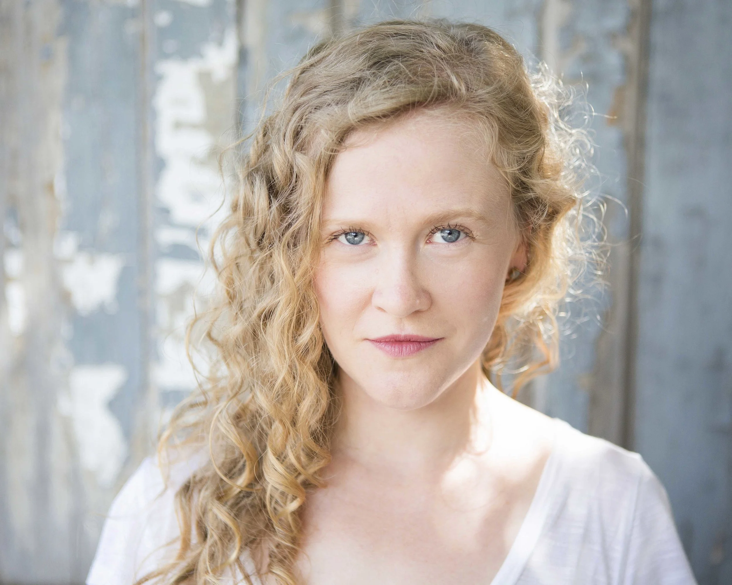 Close-up of a woman with curly strawberry blonde hair and blue eyes, wearing a white shirt, standing in front of a weathered wooden background.
