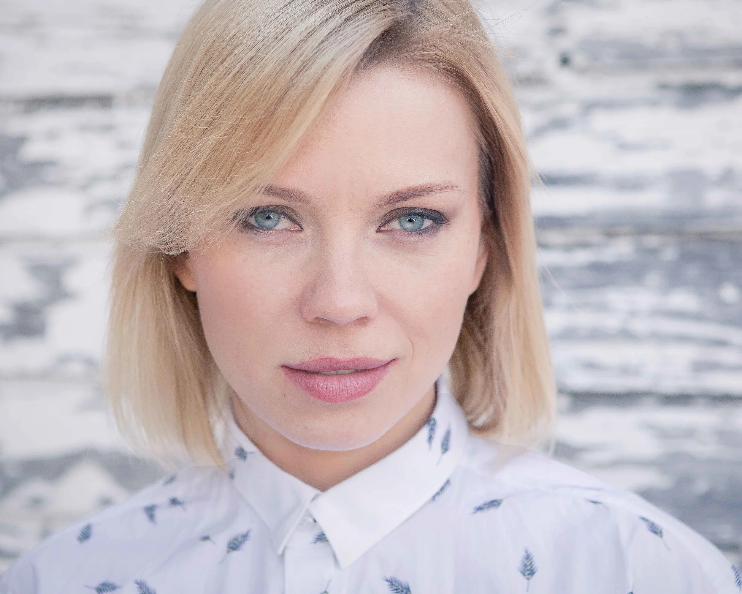 A close-up portrait of a blonde woman with blue eyes, wearing a white shirt with a small dark feather pattern, standing outdoors near water.