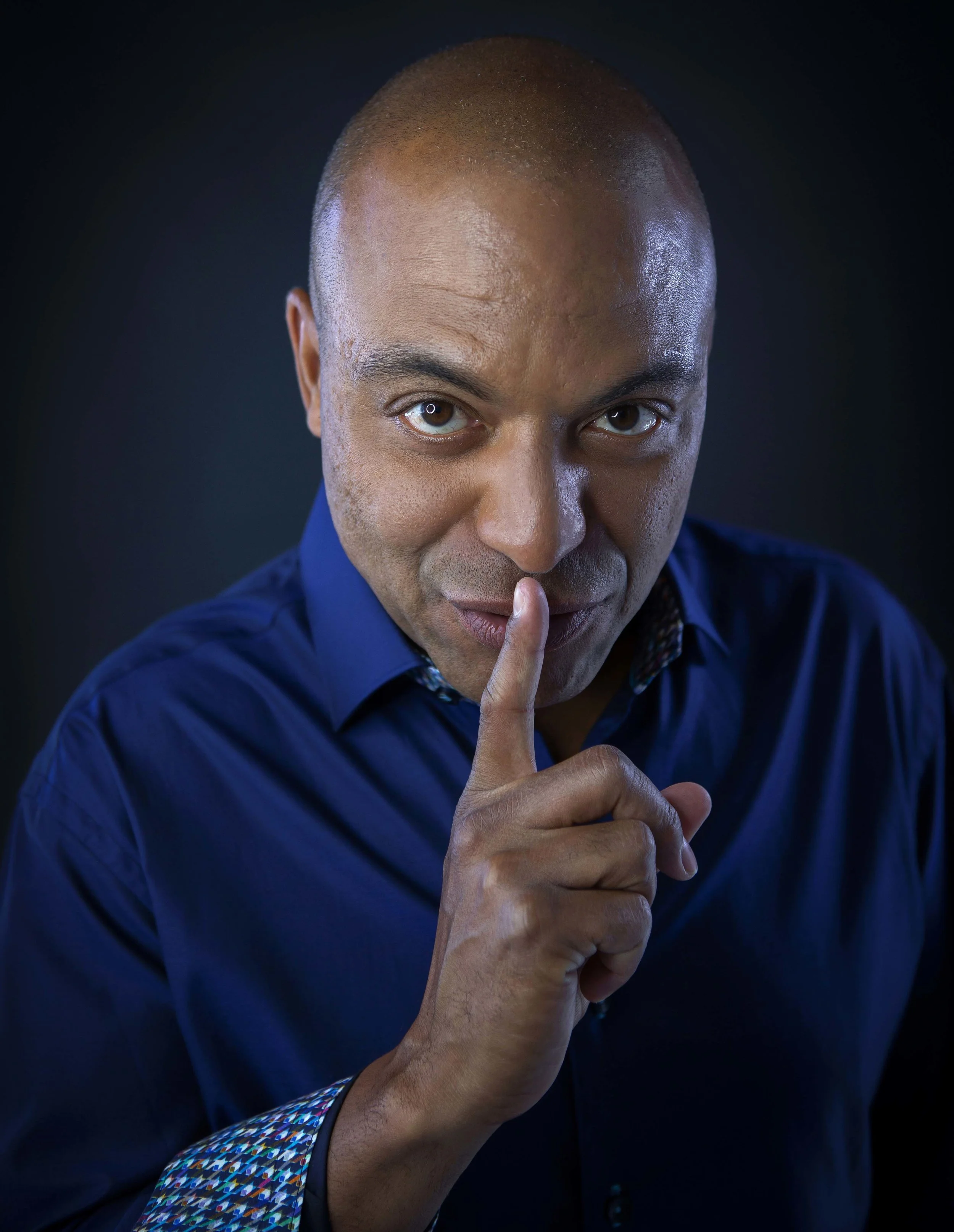 Man with a shaved head wearing a dark blue shirt, holding a finger to his lips in a shushing gesture, against a dark background.