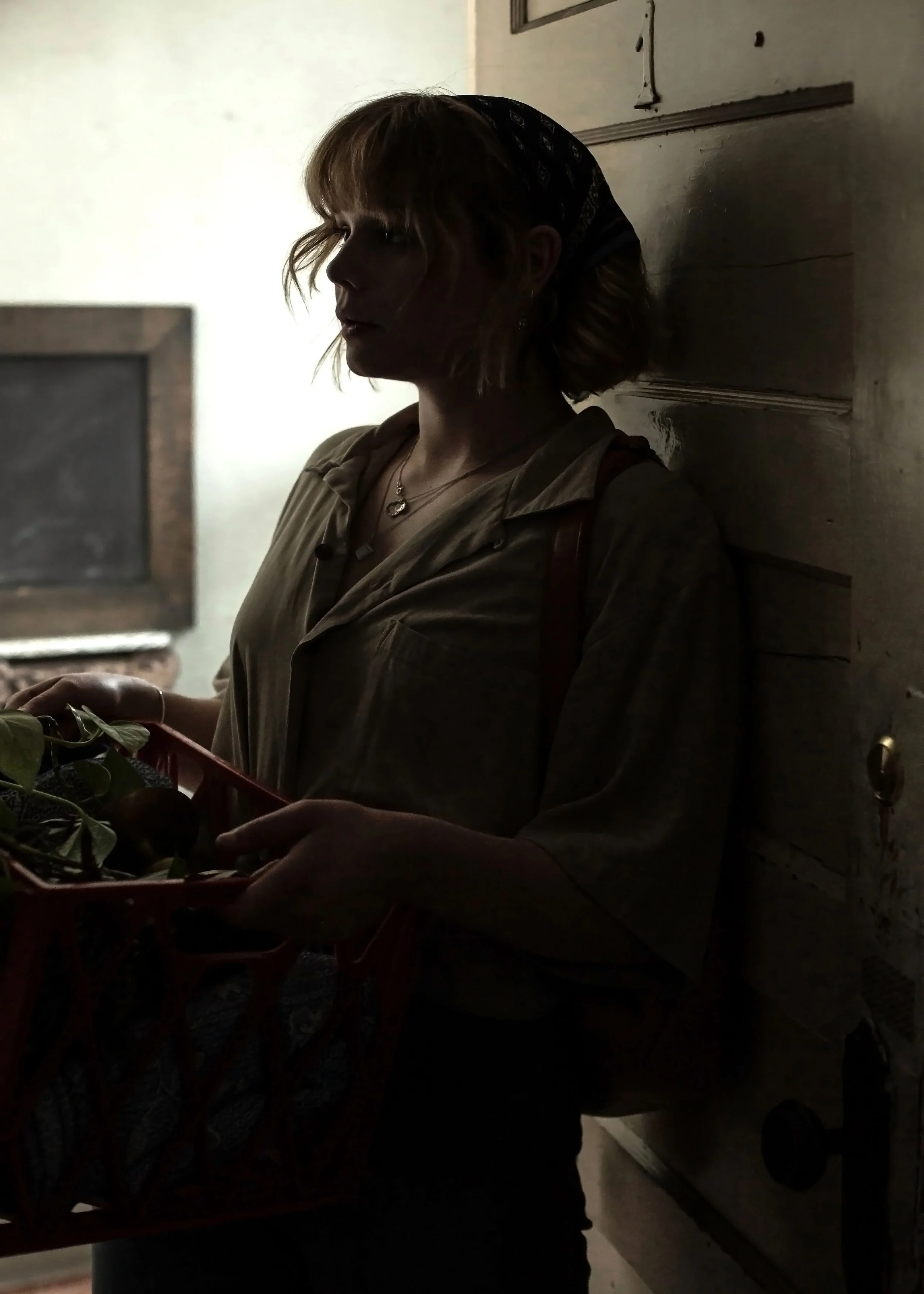 A woman standing indoors near a wall, holding a basket with greenery, and with her face in shadow.