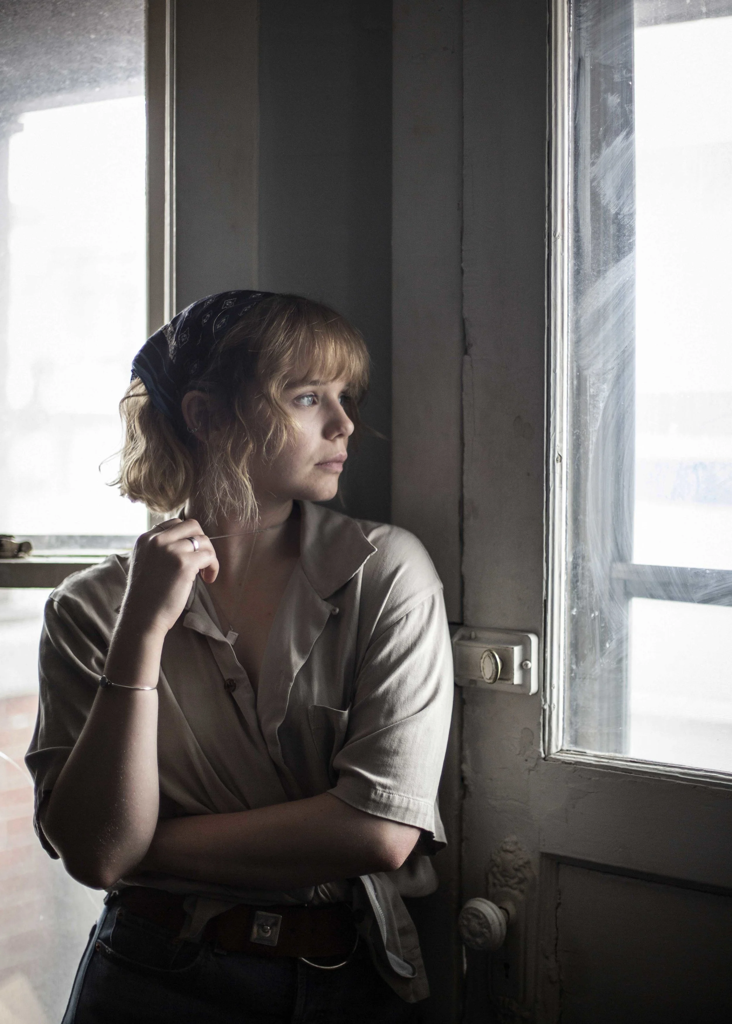 A young woman with wavy blonde hair, wearing a beige shirt and a blue bandana, stands near a window with sunlight illuminating her face, looking pensively outside.