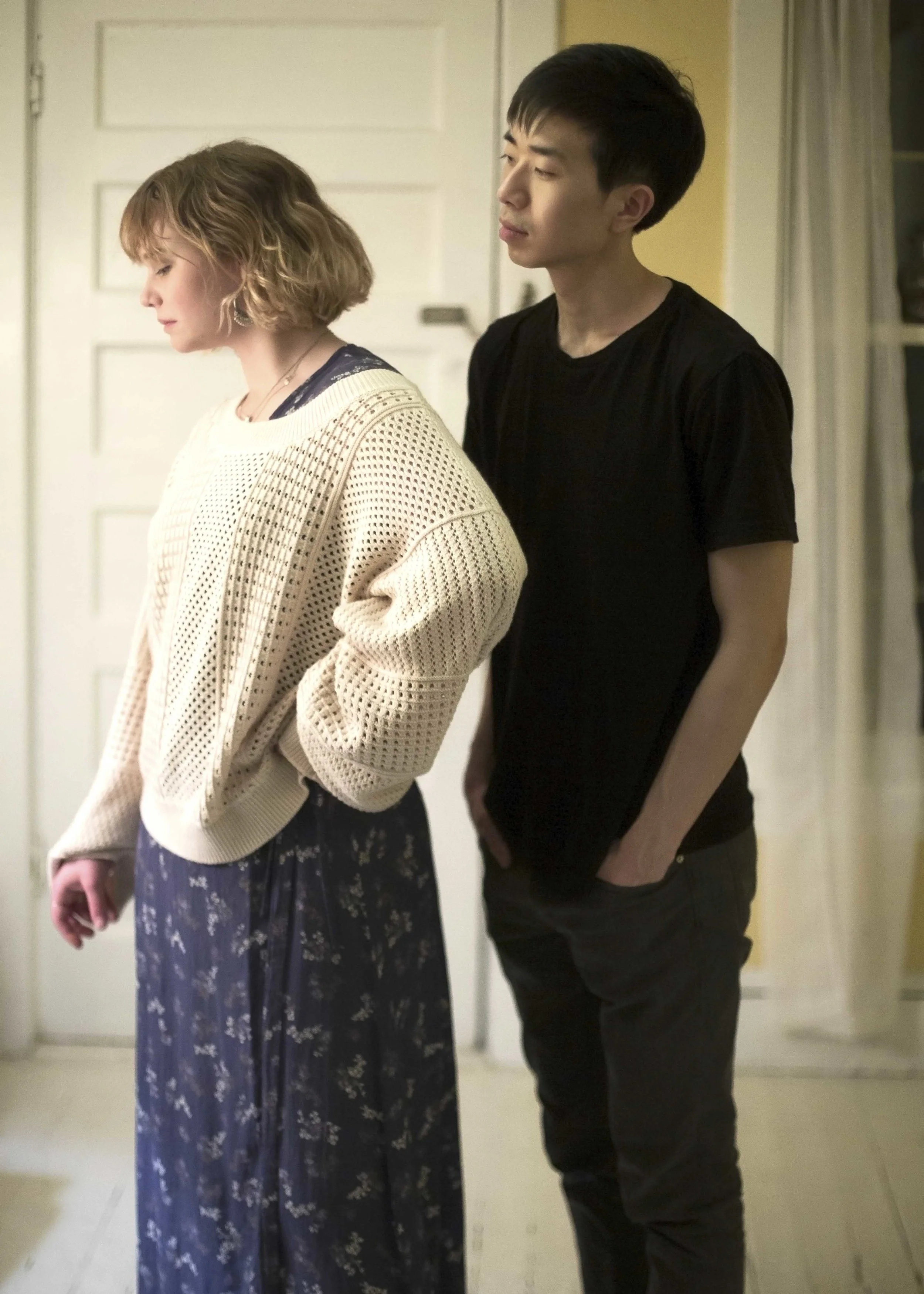 A young woman with wavy blonde hair looking down, wearing a cream-colored knitted sweater and a floral skirt, standing next to a young man with short black hair in a black shirt with his hands in his pockets, inside a room with a door and curtains in