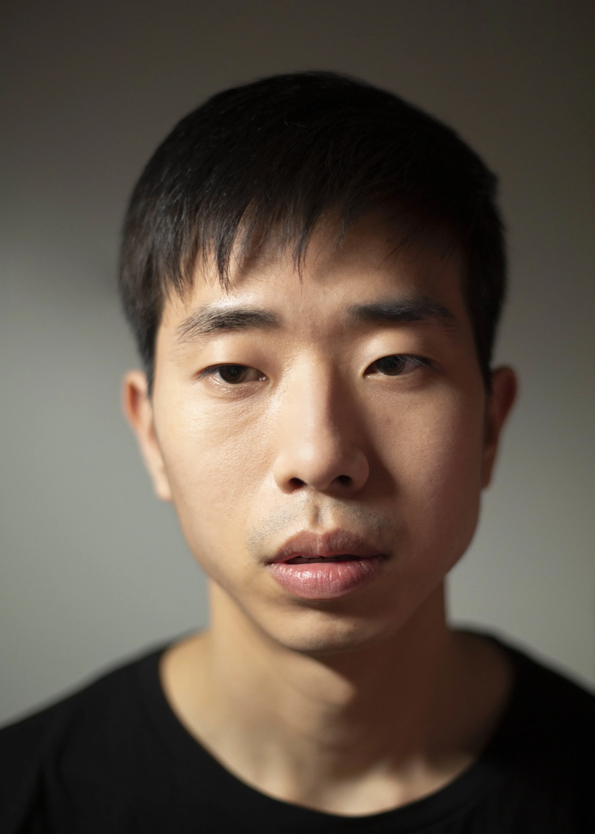 Close-up portrait of a young Asian man with short black hair, wearing a black shirt, looking slightly to the side.