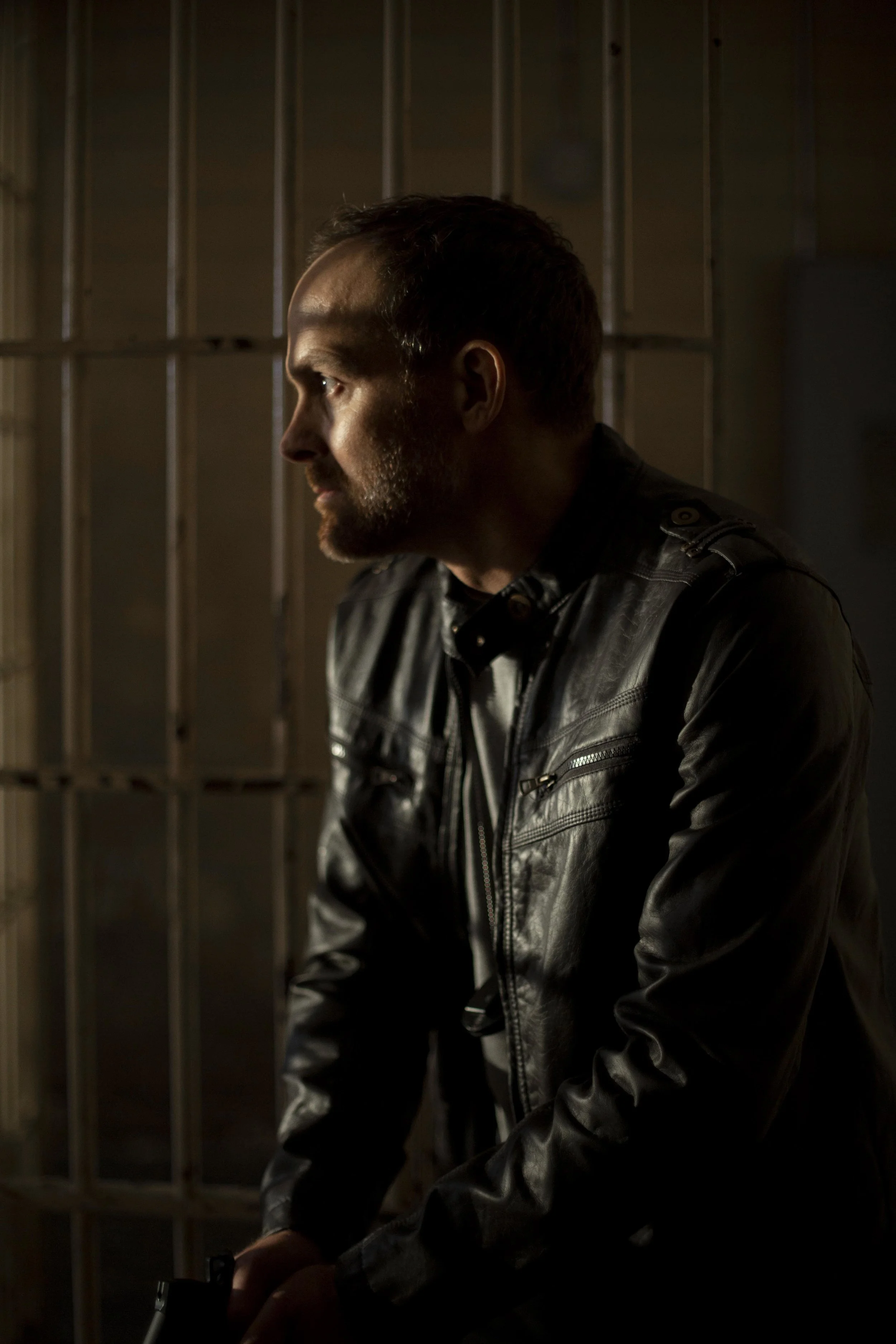 A man with a beard and short hair, wearing a black leather jacket, sitting in a dimly lit room with metal bars in the background.