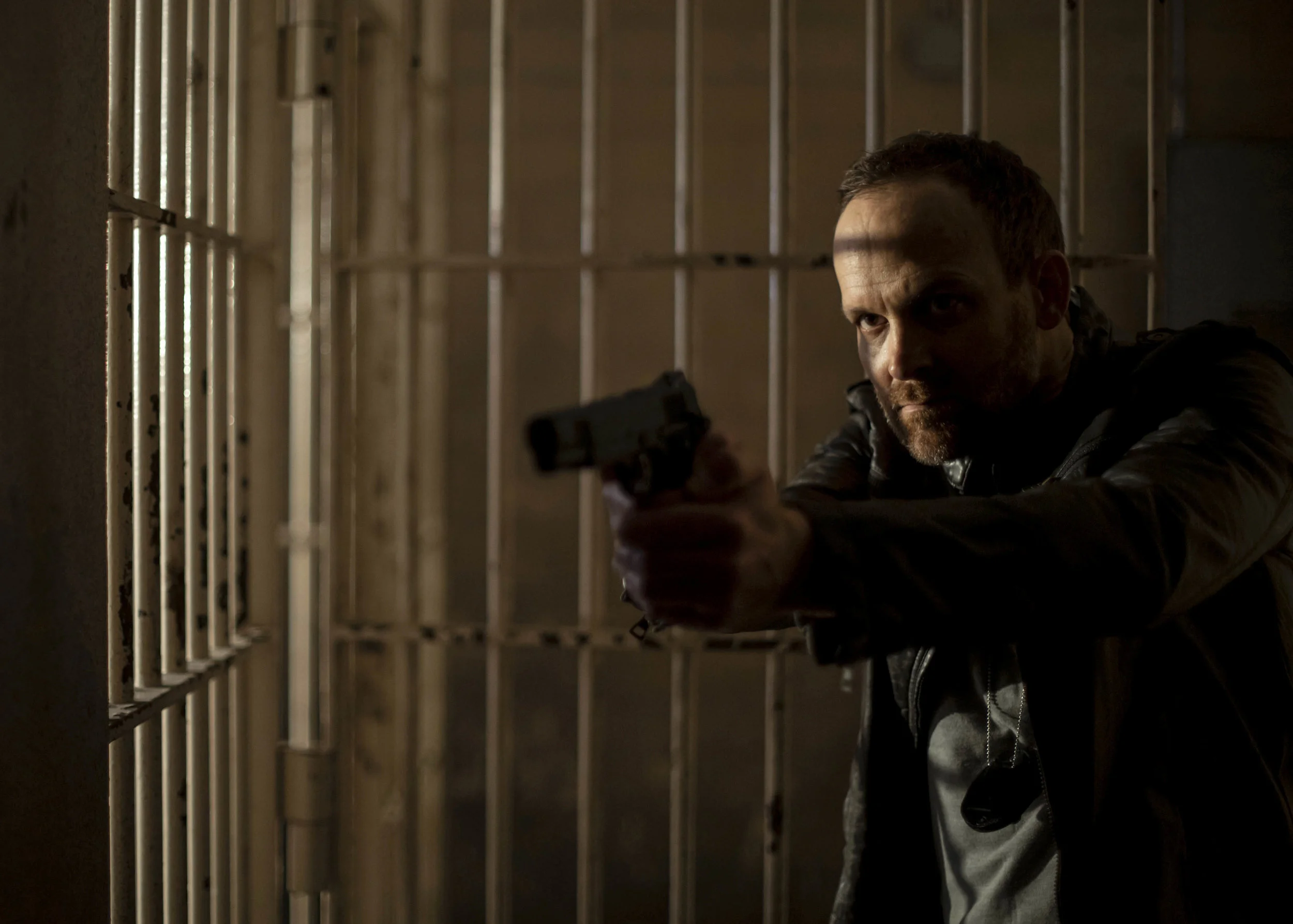 A man with a beard and short hair aiming a handgun while crouching behind metal bars in a dimly lit setting.