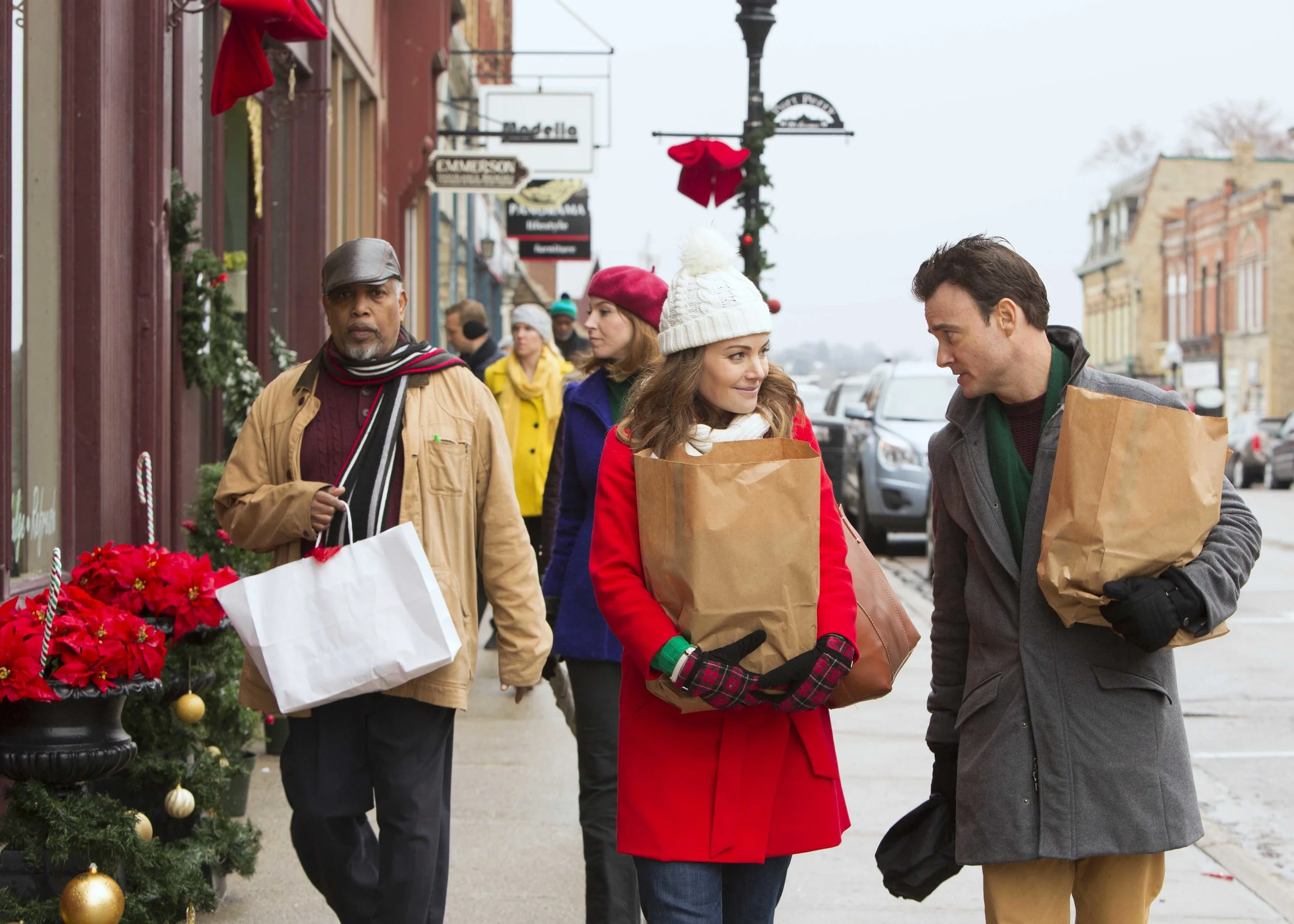 People walking on a city sidewalk dressed in winter clothing carrying shopping bags, with holiday decorations on the buildings.