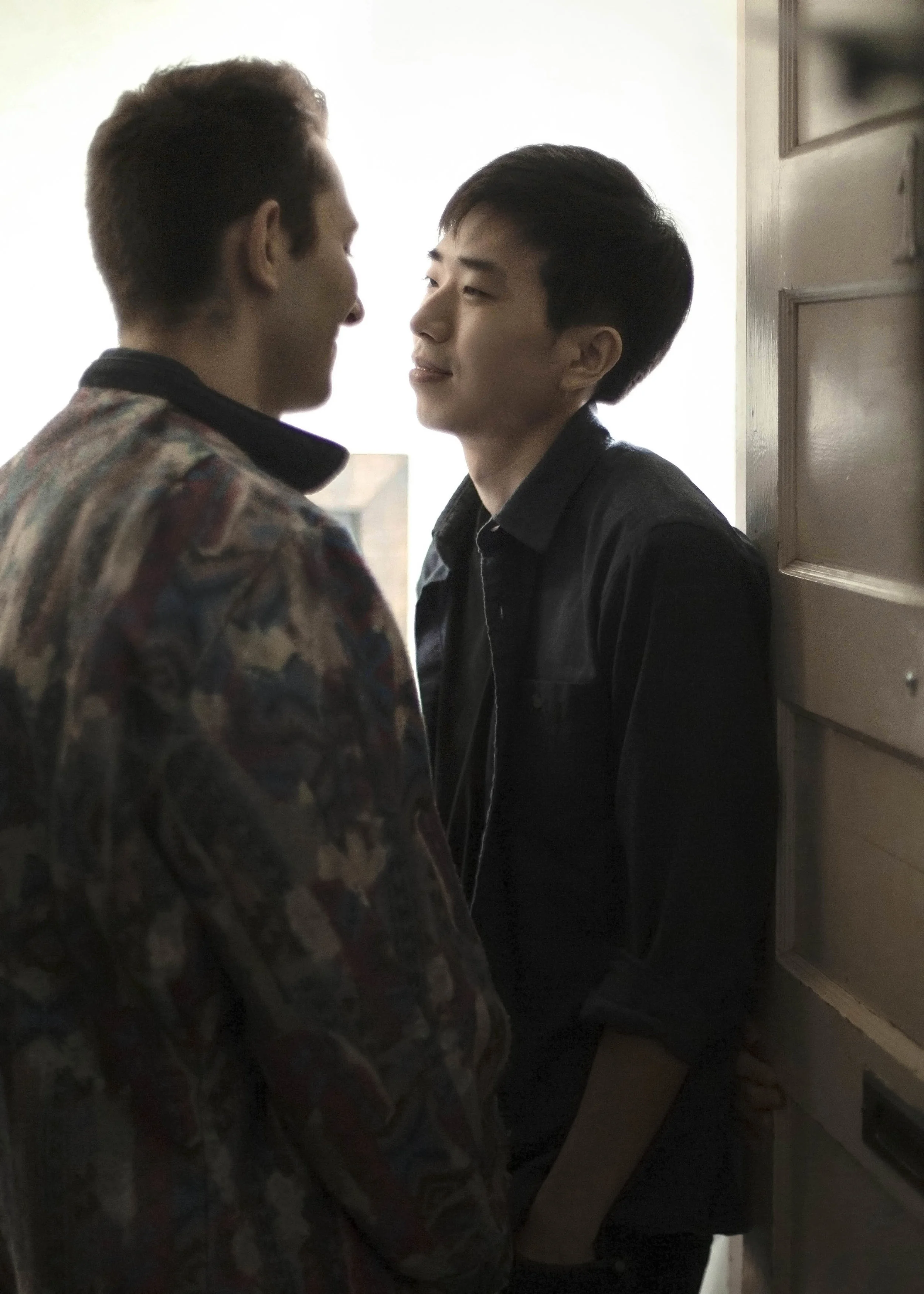Two young men standing close to each other indoors, facing each other, one with darker hair and black clothing, the other with lighter hair and multicolored patterned jacket, engaging in an intense or intimate moment.