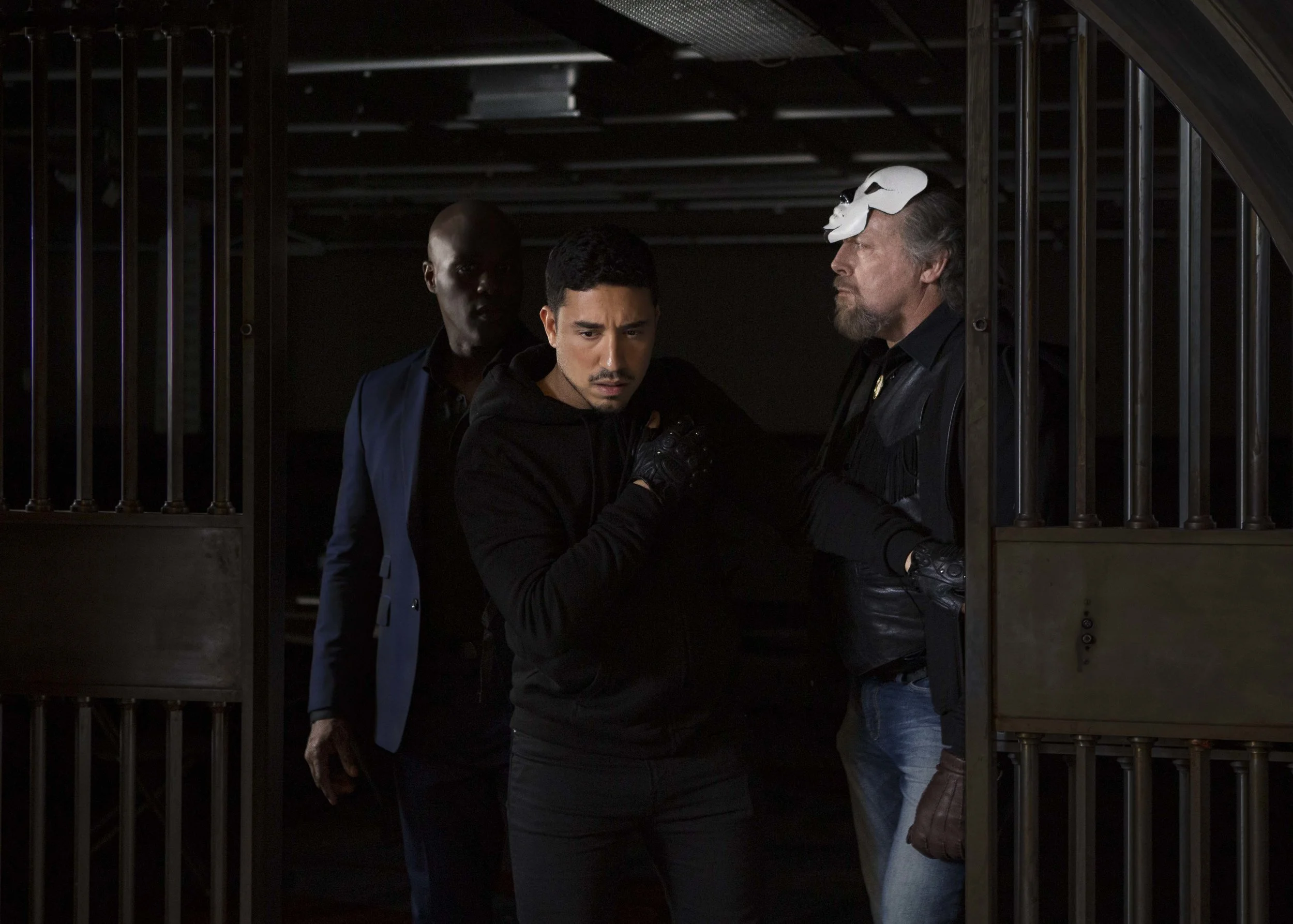 Three men walking through a dark, industrial-looking corridor with metal bars, one of them wearing a white mask on his head.