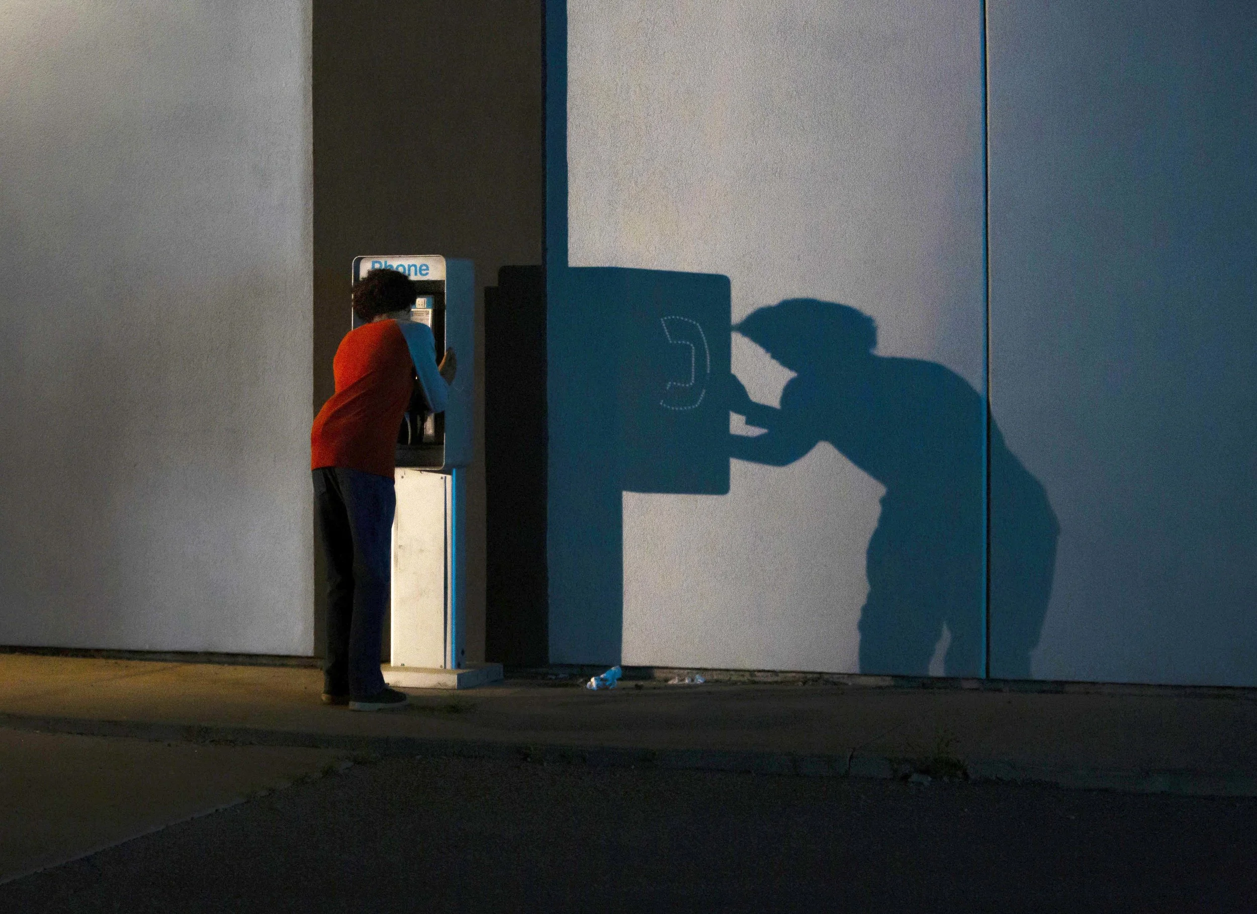 A young boy at a payphone booth on a dark street, casting a shadow that resembles a person using the phone.
