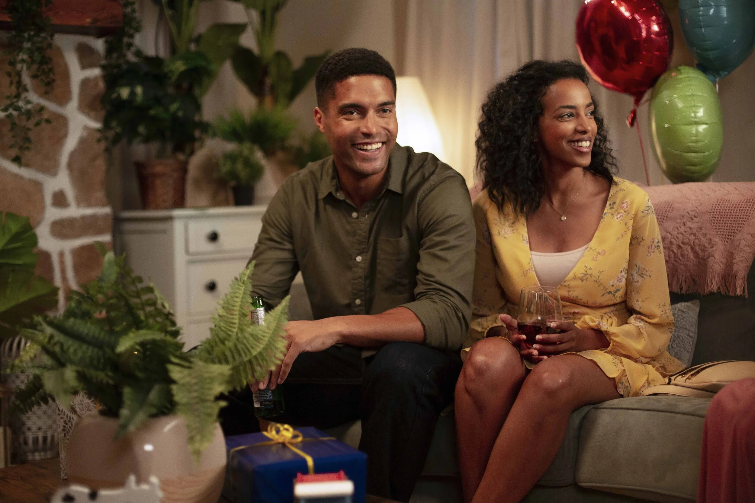 A happy man and woman sitting on a couch at a party, with balloons and gifts around them, holding a glass of wine.