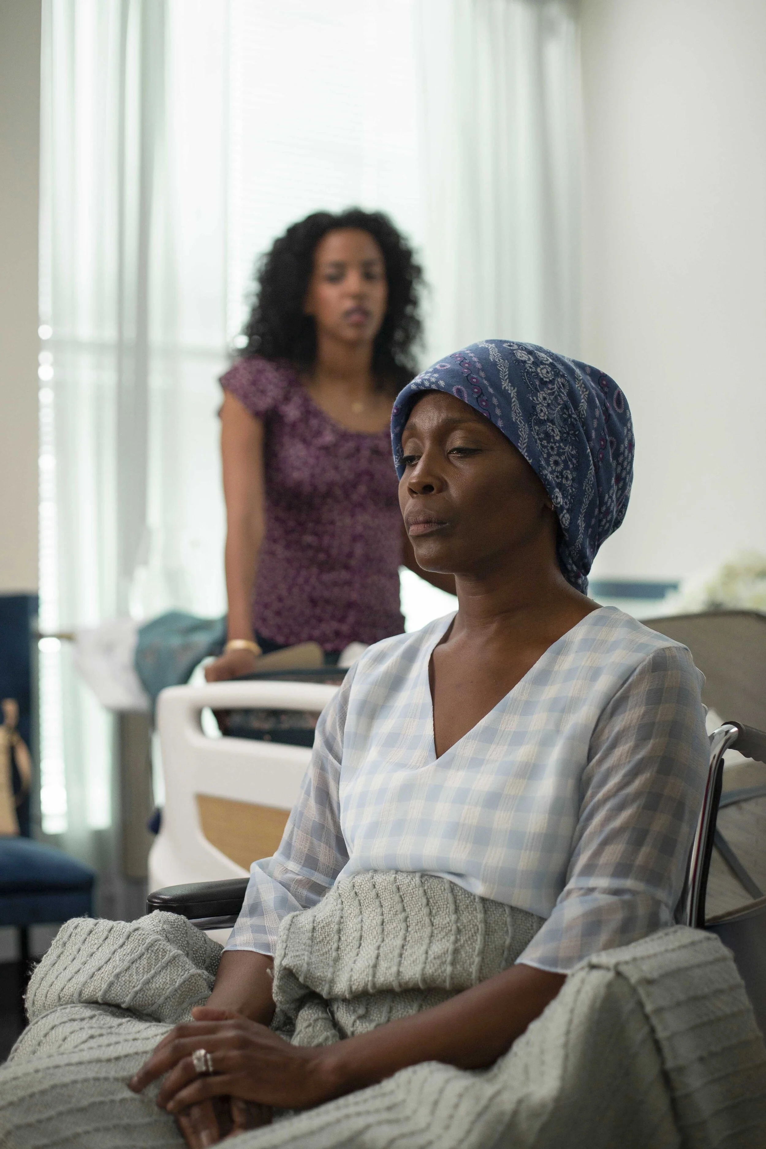 A woman in a hospital bed wearing a headscarf, with a blanket on her lap, looks somber. A nurse or visitor stands in the background, blurred, in a hospital room with curtains and medical equipment.