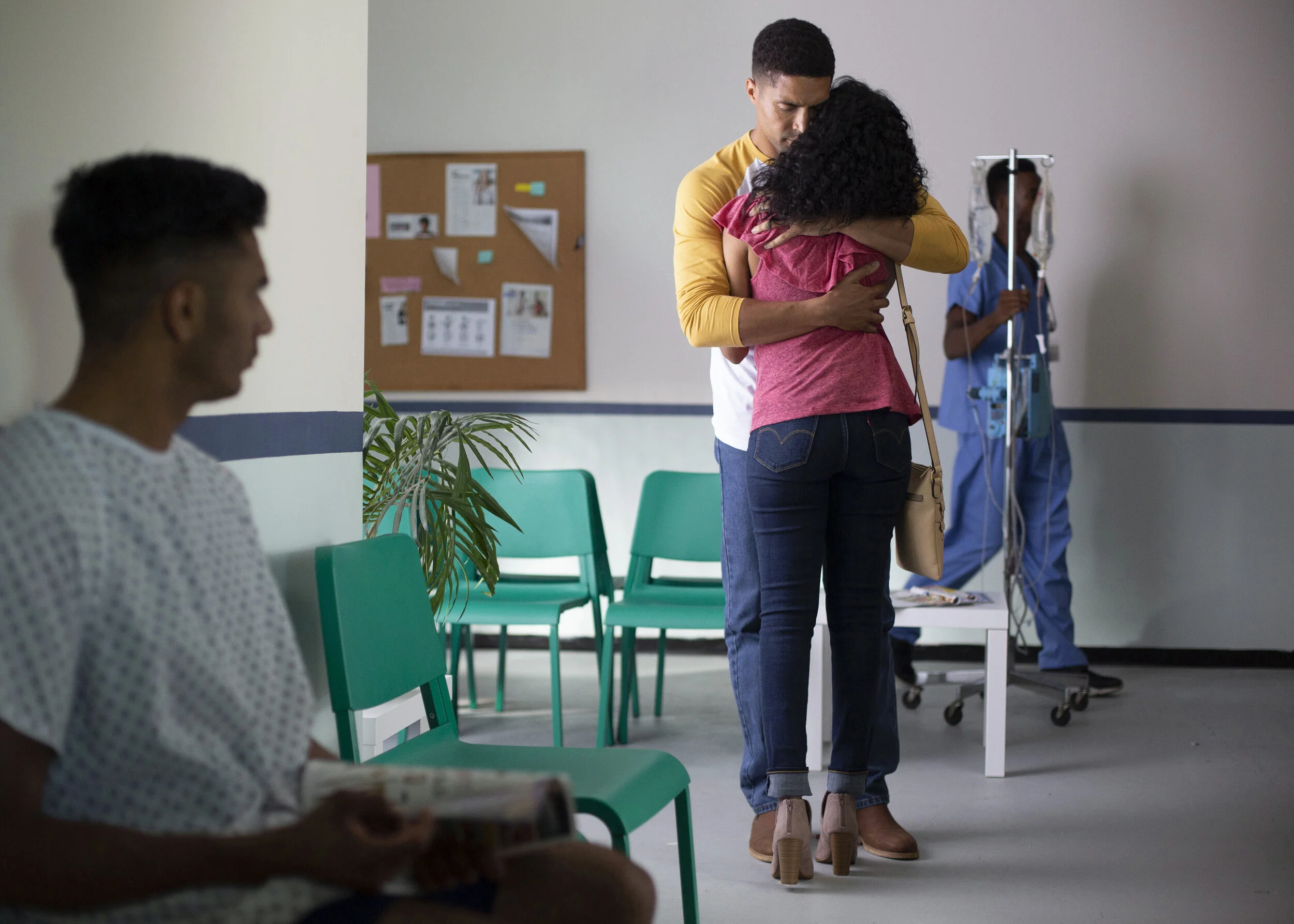 A couple embraces in a hospital waiting room while a man in a hospital gown waits nearby, and a nurse stands in the background holding an IV pole.