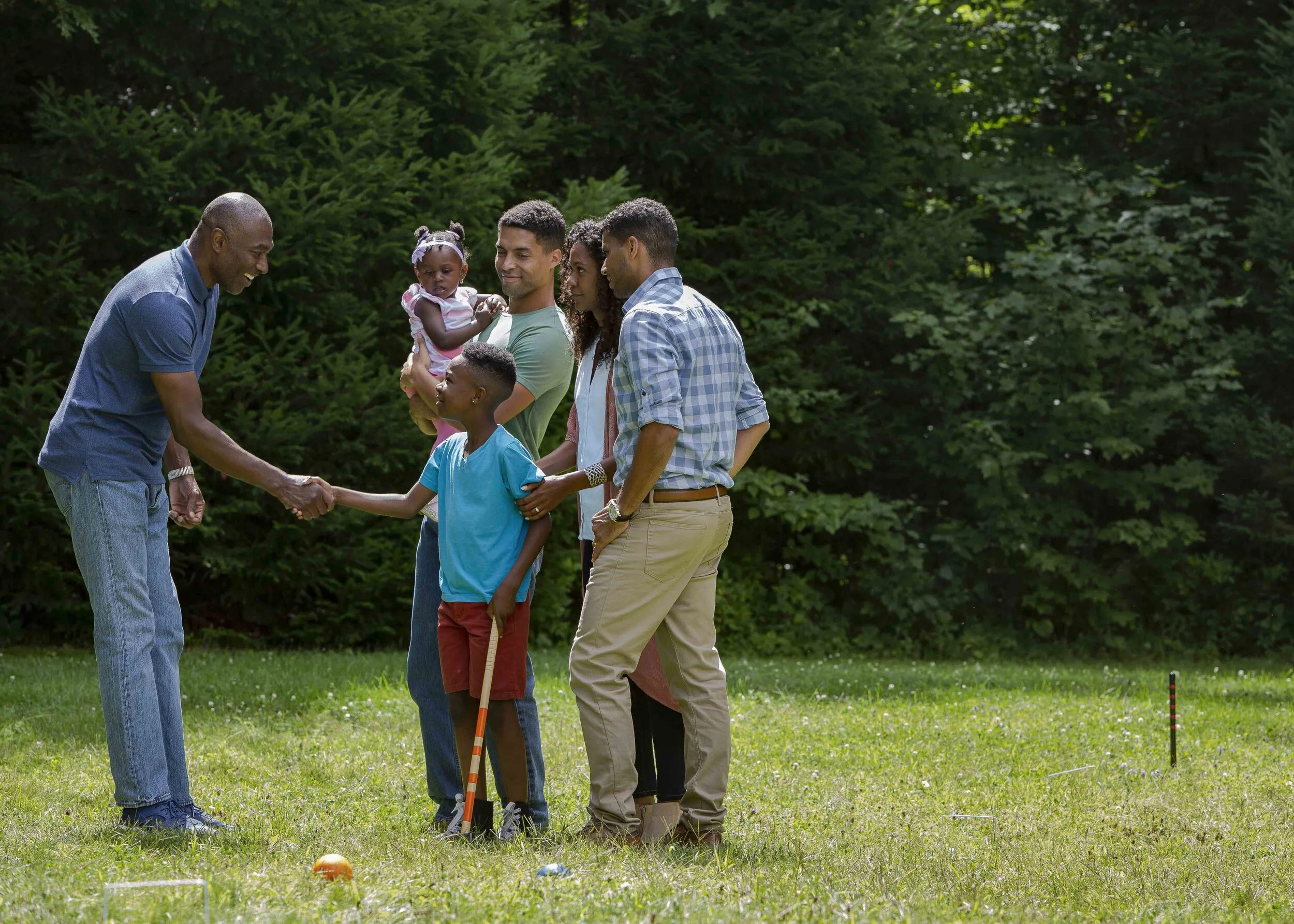 A group of people, including a man in a blue shirt shaking hands with a young boy, standing on a grassy field in a park with trees in the background.