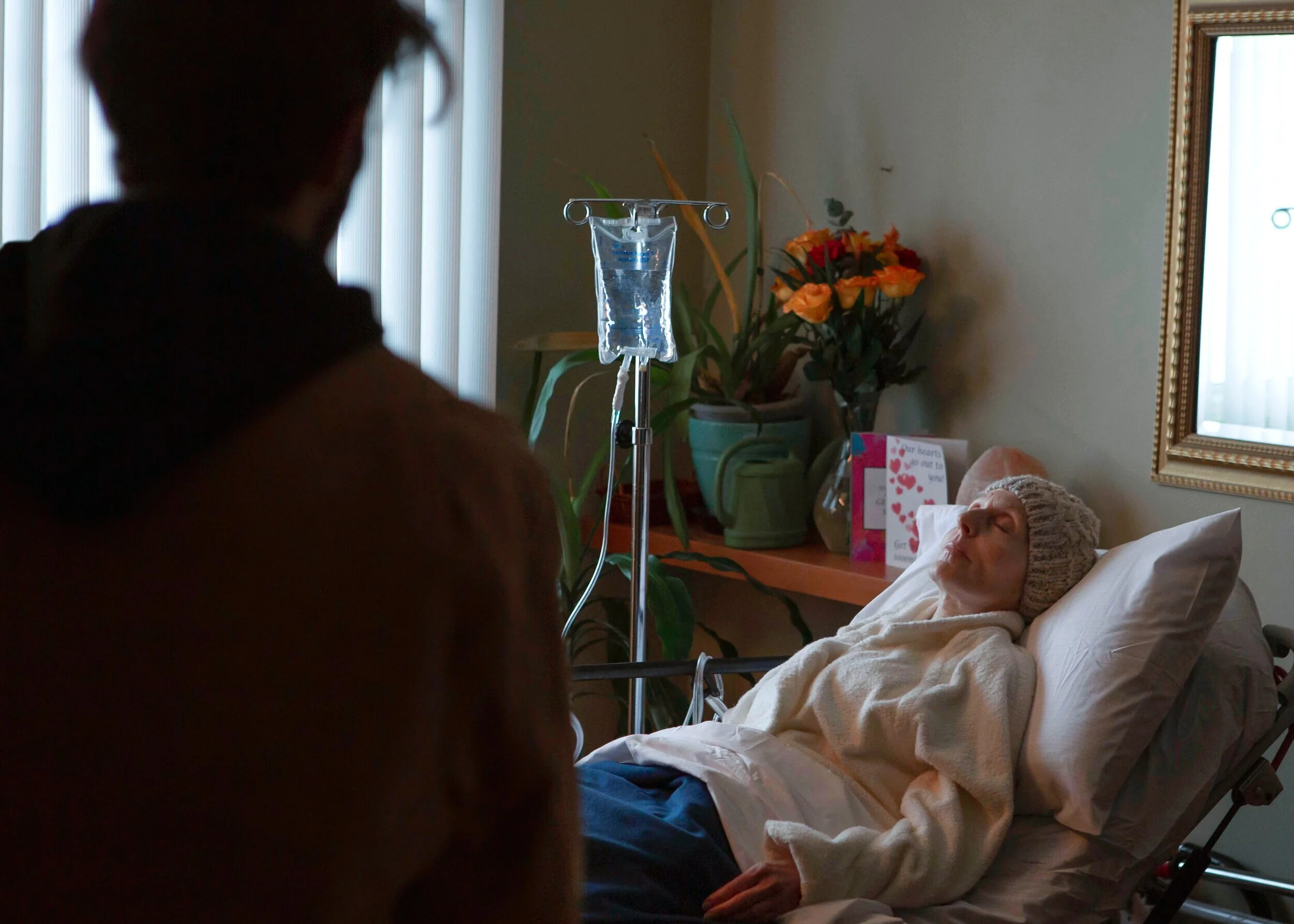 An elderly woman in a hospital bed with a knit cap, appearing weak, is lying under a blanket. A person is standing nearby, blurred in the foreground. There is an IV drip next to her, and potted plants and a greeting card are visible on a table behind