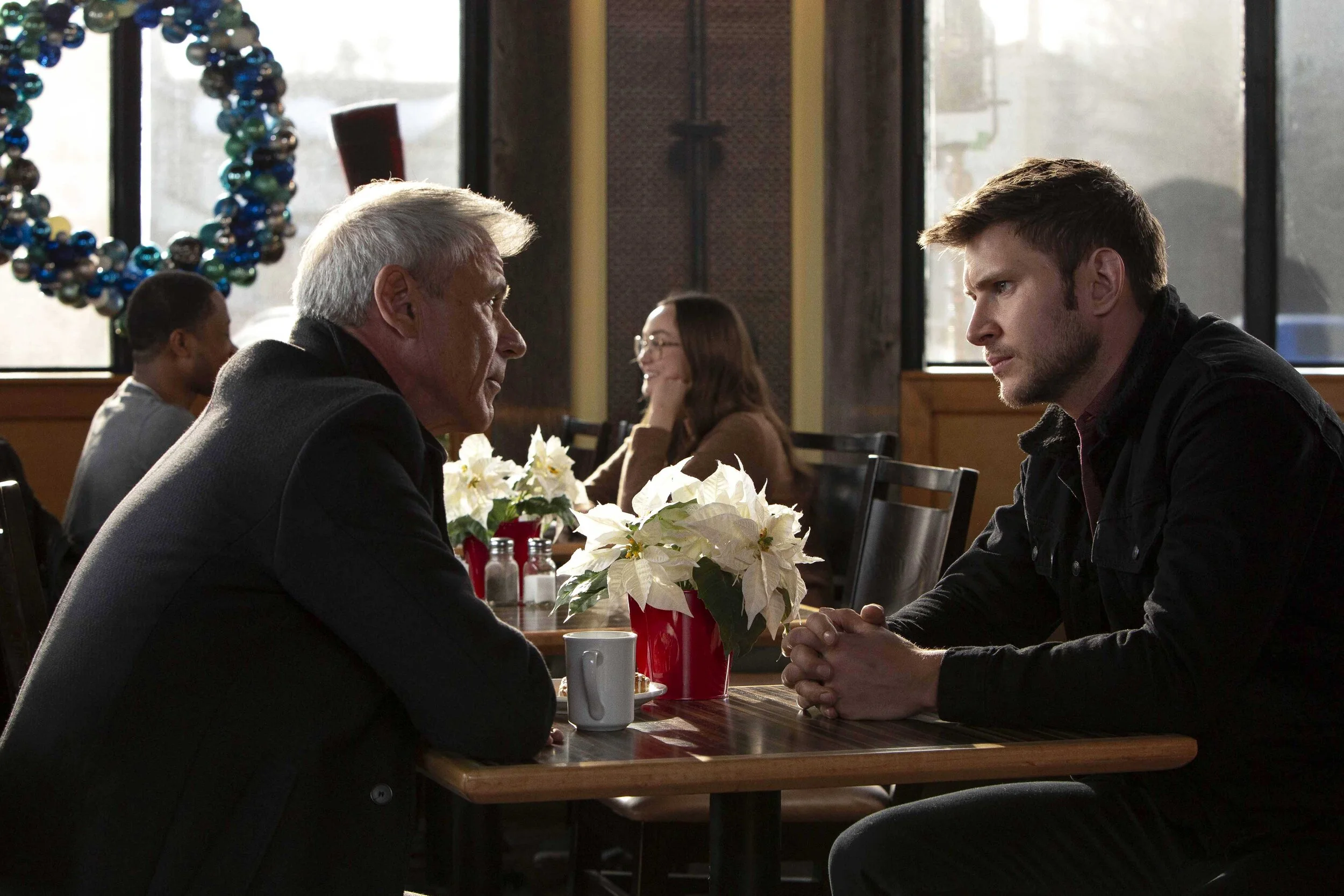 Two men sitting across from each other at a restaurant table, engaged in conversation. The older man has gray hair and is wearing a black jacket, while the younger man has brown hair and is also wearing dark clothing. There are white poinsettia flowe