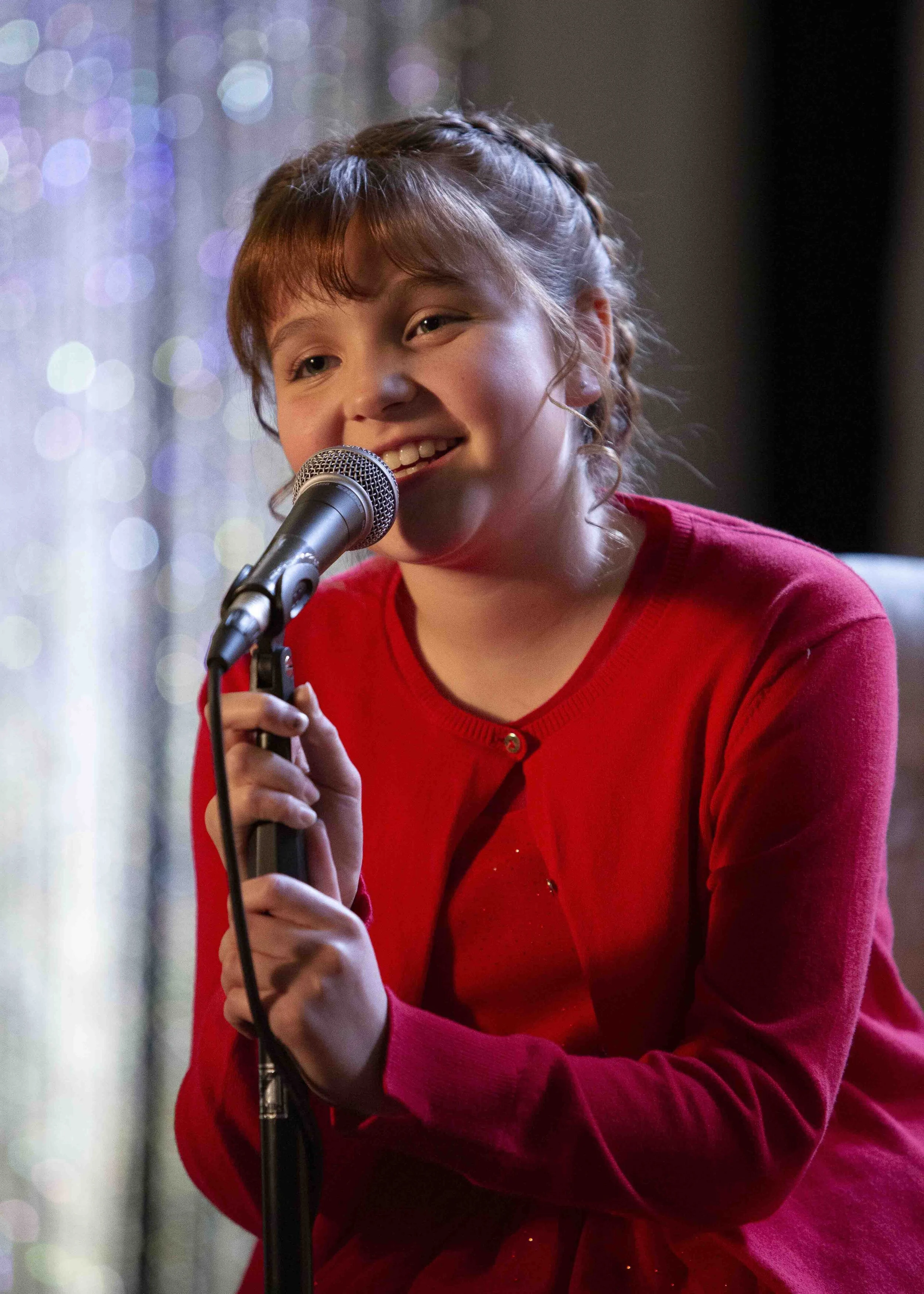 A young girl singing or speaking into a microphone, wearing a red cardigan and smiling, with colorful lights in the background.