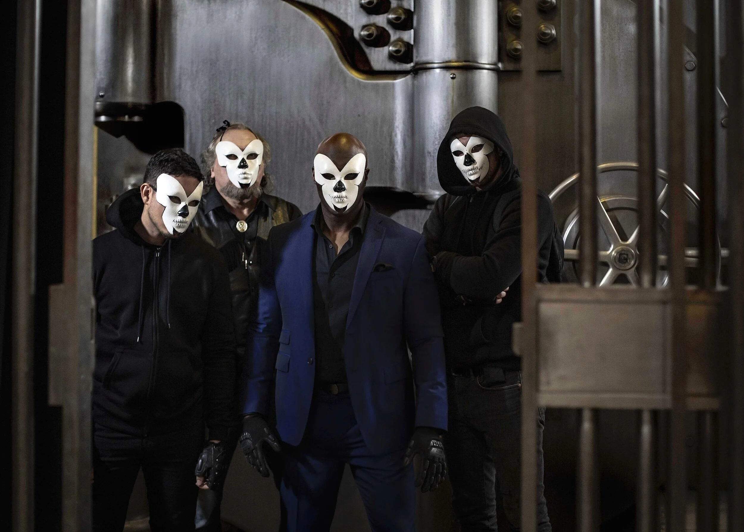 Five individuals wearing white skull masks, four of them in black hoodies and one in a blue suit, inside an industrial setting with metal pipes and doors.