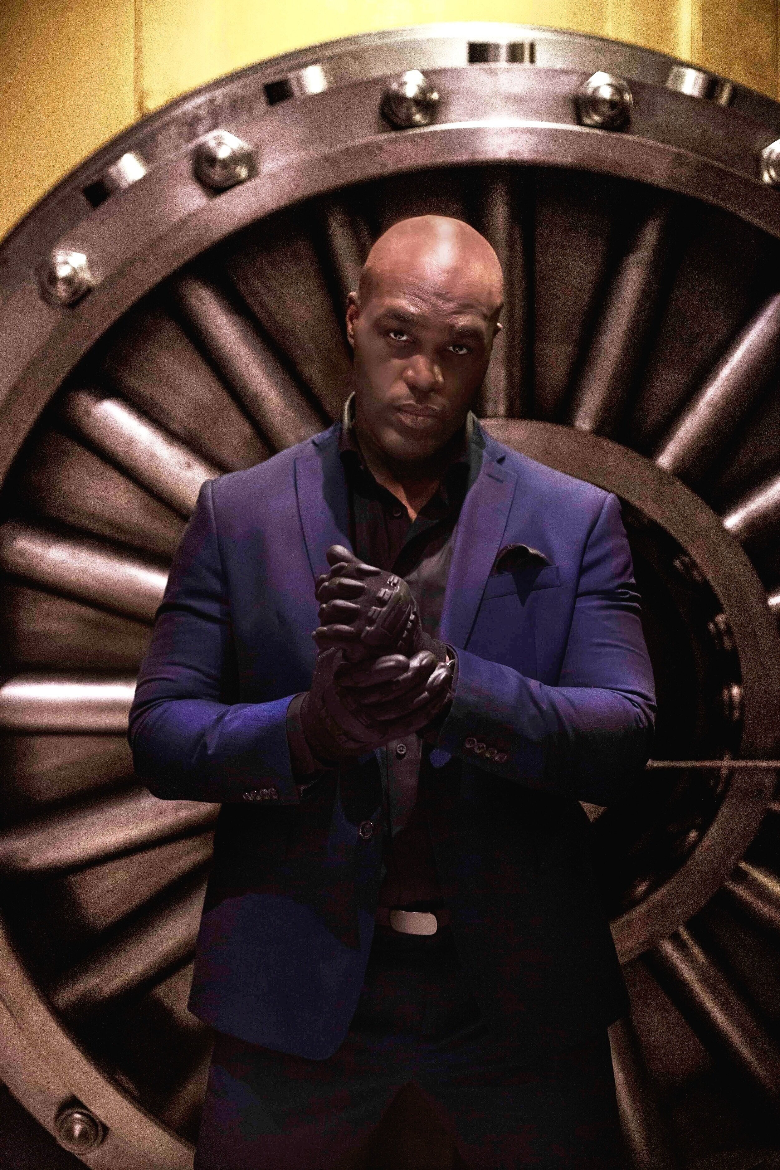 A man in a dark blue suit and black gloves stands in front of a large, circular industrial vault door. He has a serious expression and is clasping his gloved hands together.