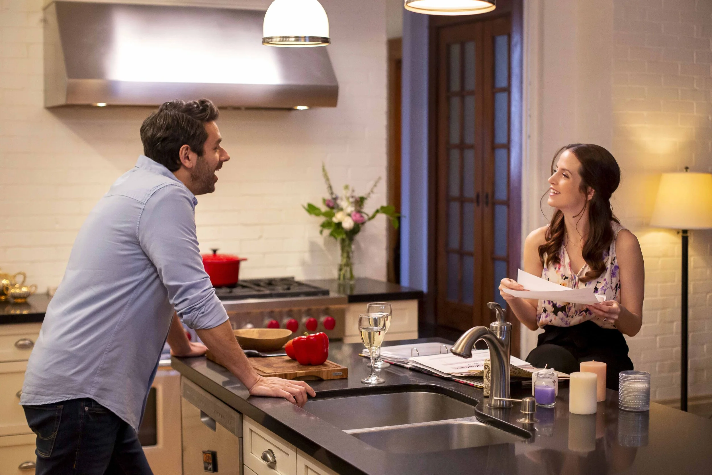 A man and a woman are having a lively conversation in a kitchen, smiling and leaning on the kitchen counter with a red pepper, a glass of white wine, and some papers in front of them.