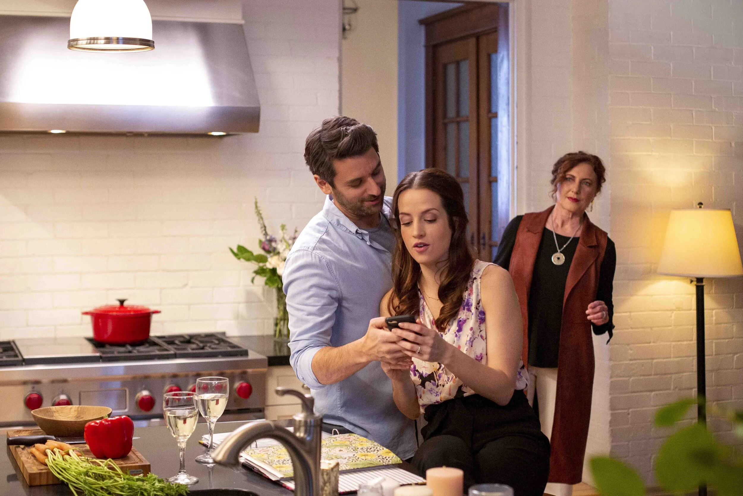 A young man and woman are standing in a kitchen, looking at a phone together, while an older woman observes from the background. The kitchen has a stove, a red pot, glasses of water, and various vegetables on the counter.