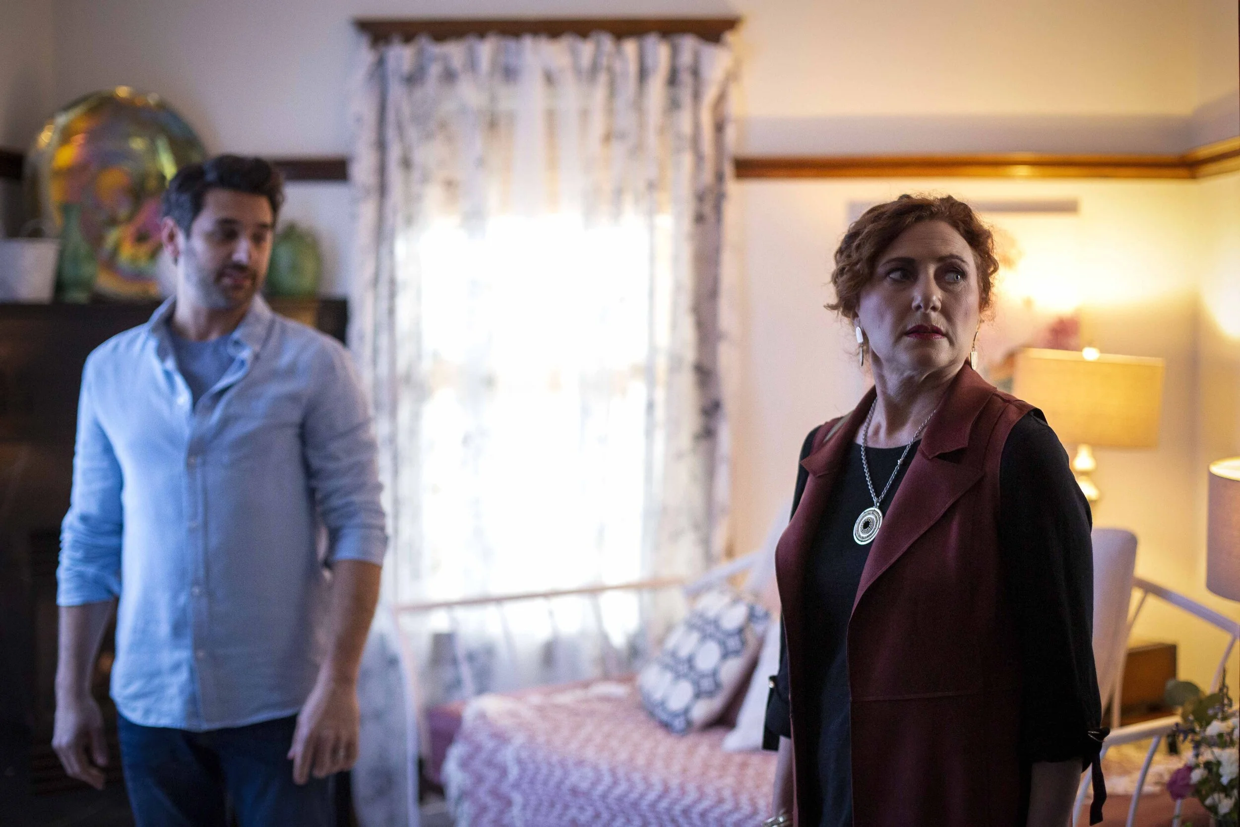 A man and a woman standing in a living room with a sofa, framed pictures, and lamps, with sunlight coming through a window.
