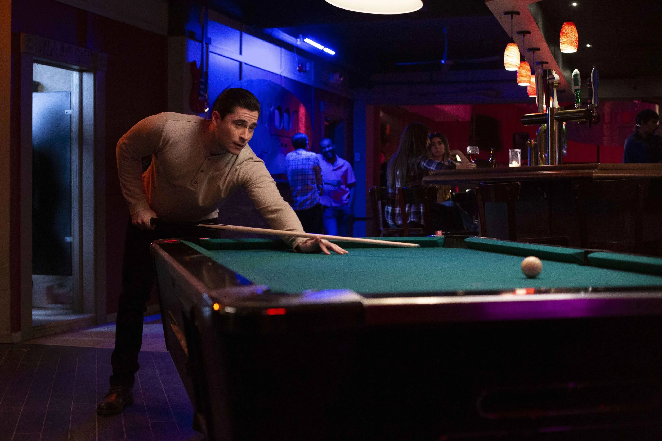 A man playing pool at a bar, preparing to take a shot, with other people sitting and standing in the background.