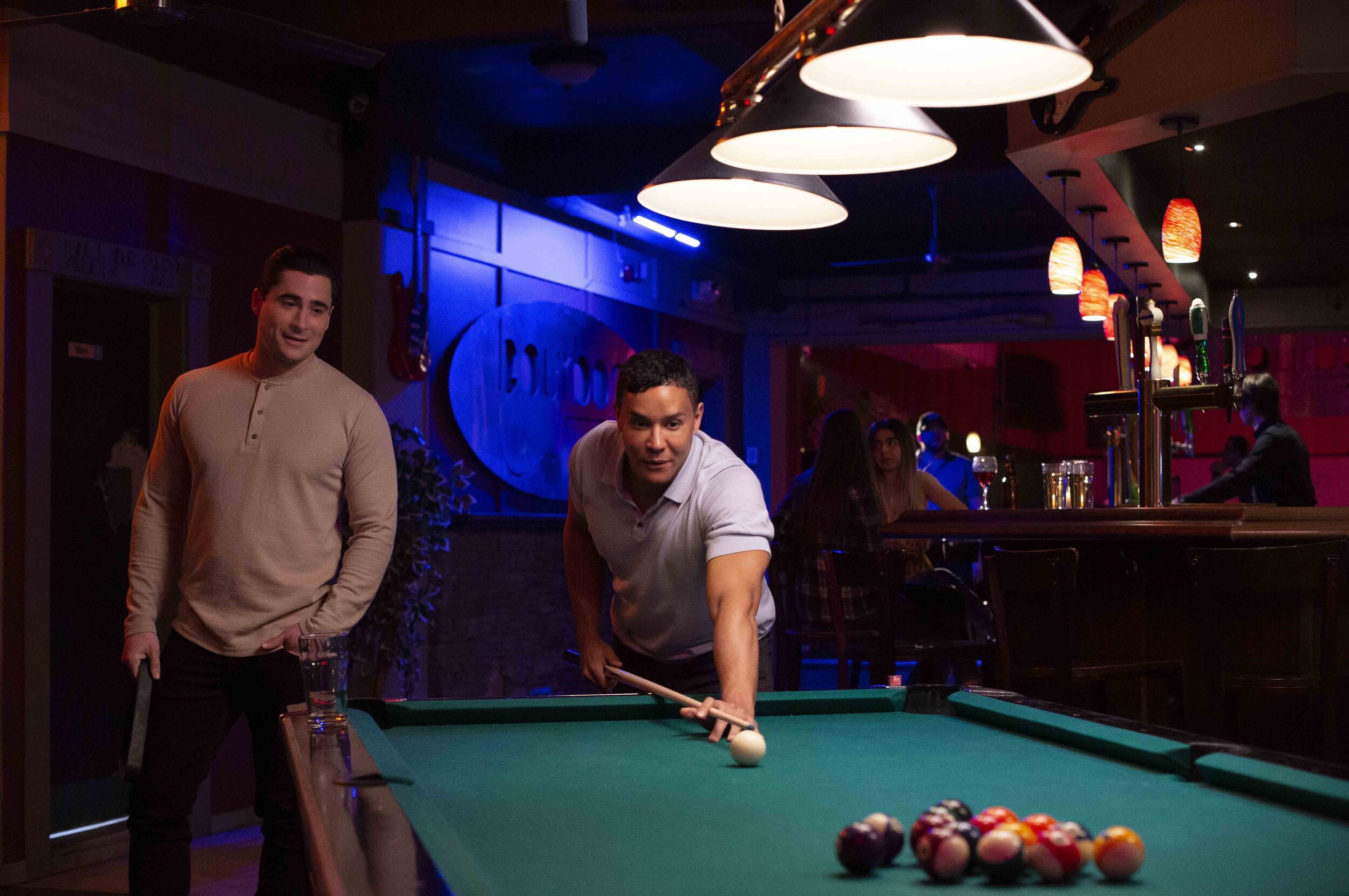 Two men playing pool at a bar, with one preparing to take a shot and the other observing. The scene is dimly lit with bar counter and hanging lamps overhead, and there are other patrons in the background.