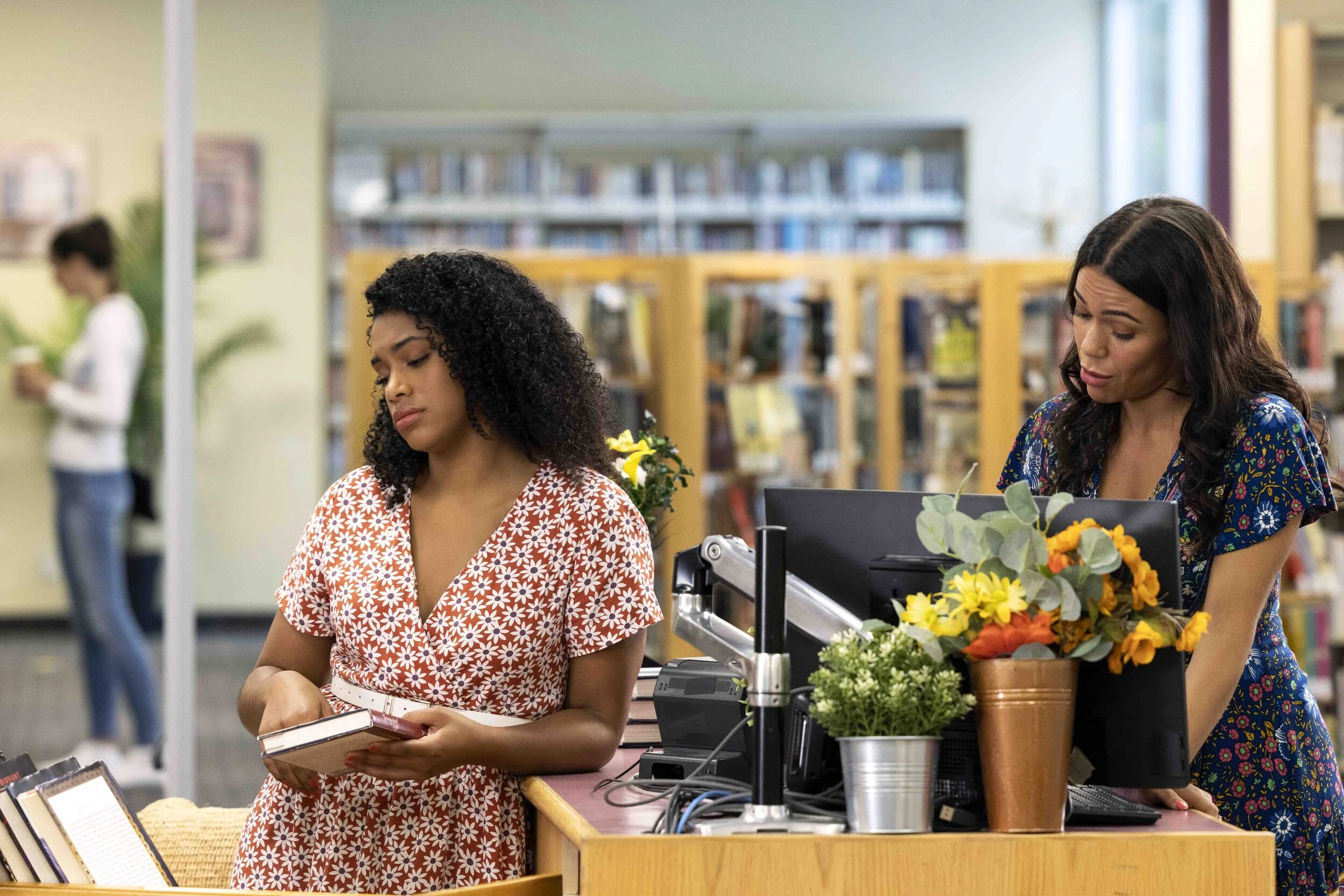 A woman in a red and white floral dress looks at her phone in a bookstore. Another woman in a blue patterned dress is behind the counter, working on a computer. The counter has flowers and plants.