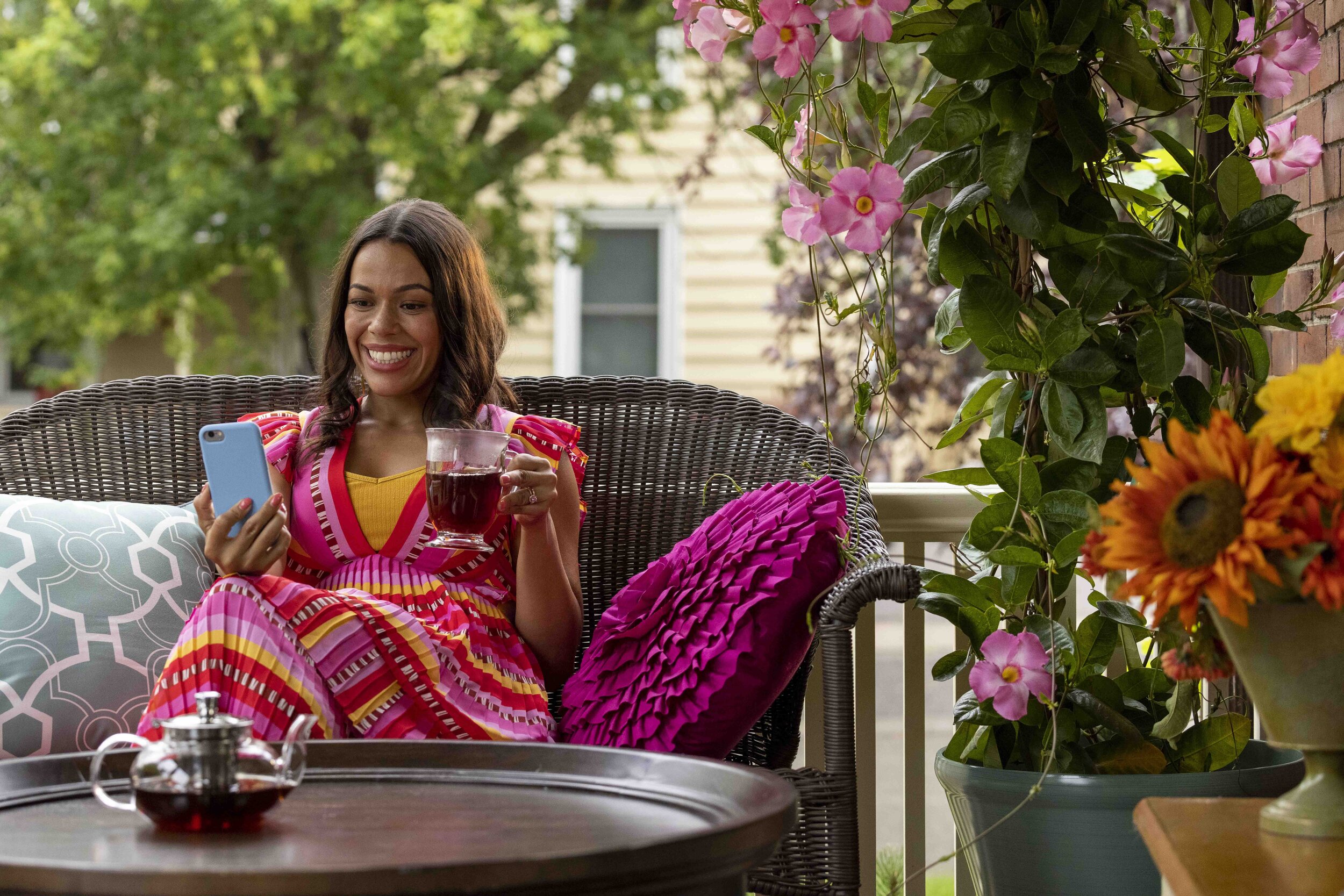 A woman sitting on a porch, wearing a colorful pink and yellow striped dress, smiling while looking at her phone and holding a glass of red wine. There are pink and yellow flowers and lush green trees in the background, with a teal pillow with a geom