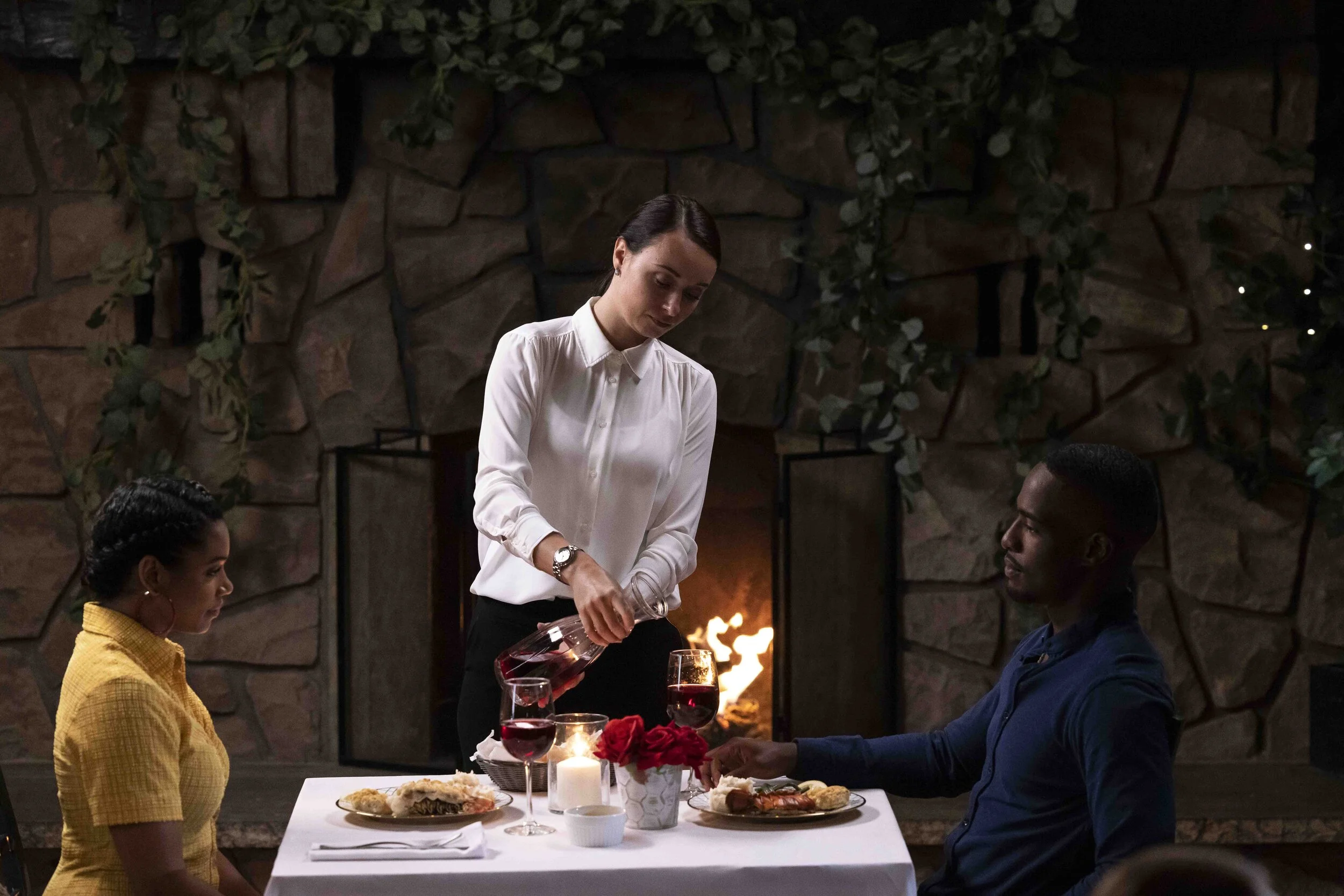 A woman pouring red wine into a glass at a dinner table with two seated guests, one man and one woman, in a cozy restaurant with a stone fireplace and green foliage in the background.
