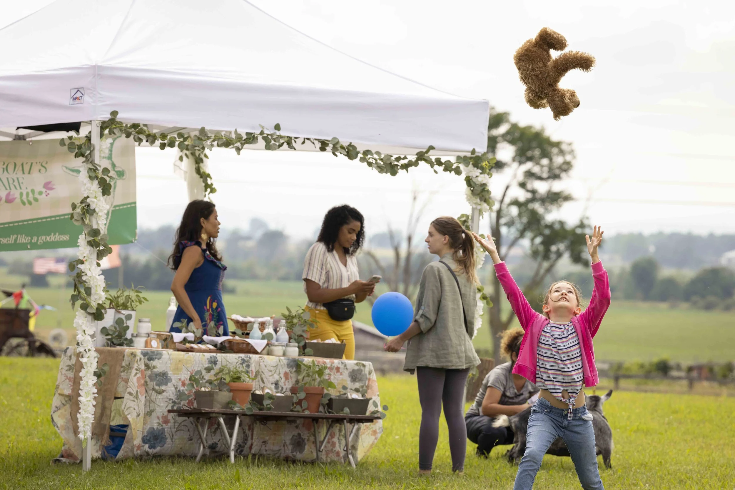 A young girl in a pink jacket and striped shirt throws a teddy bear into the air during an outdoor event with vendors, children, and animals in a grassy field.