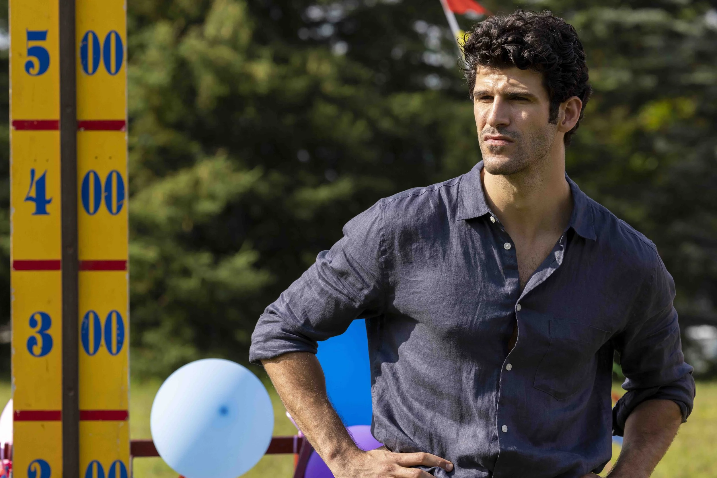 A man with curly dark hair and a serious expression standing outdoors next to a yellow measuring stick with blue numbers, with balloons in the background.
