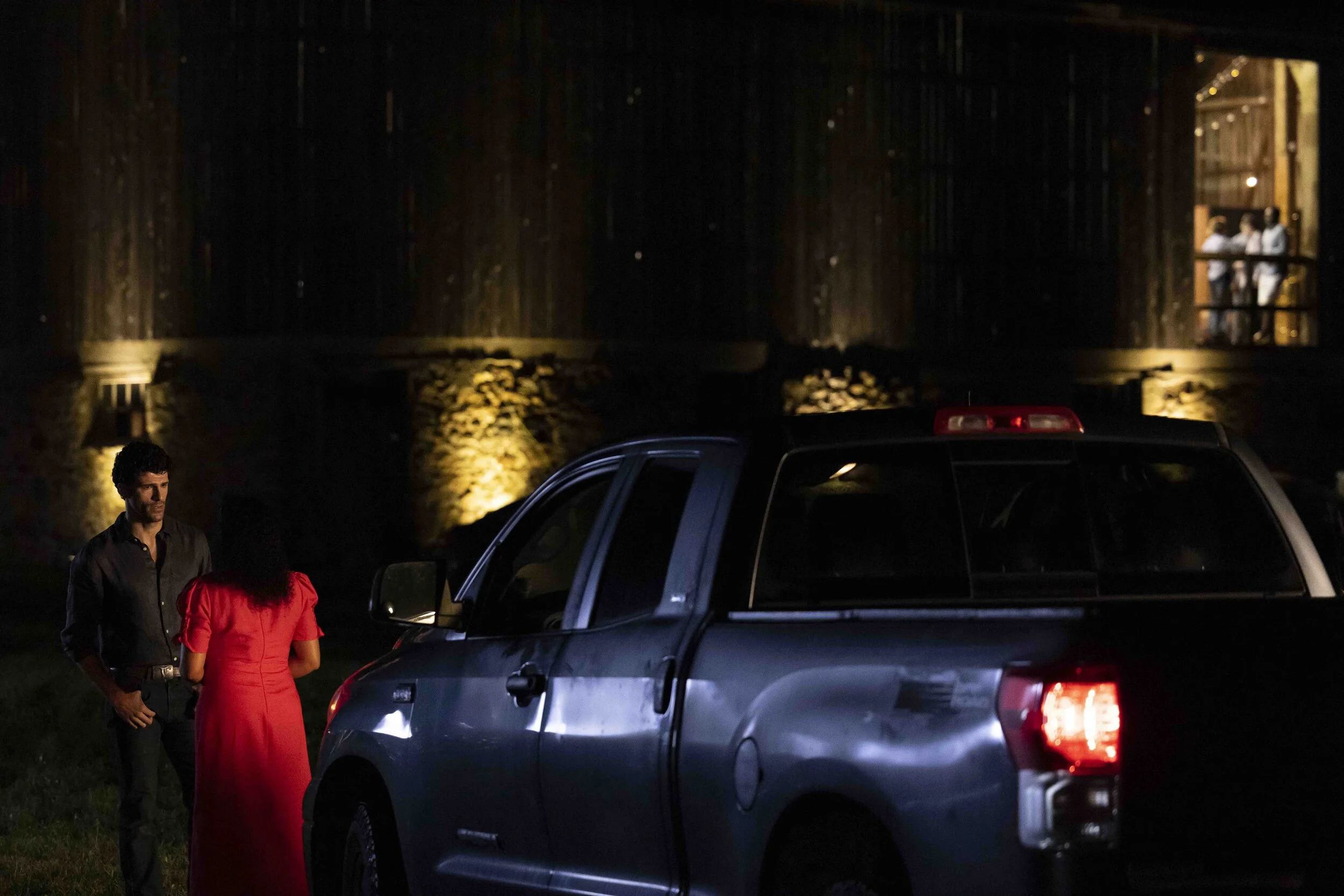 A man and a woman standing beside a pickup truck at night, with a lit building in the background and people on a balcony.