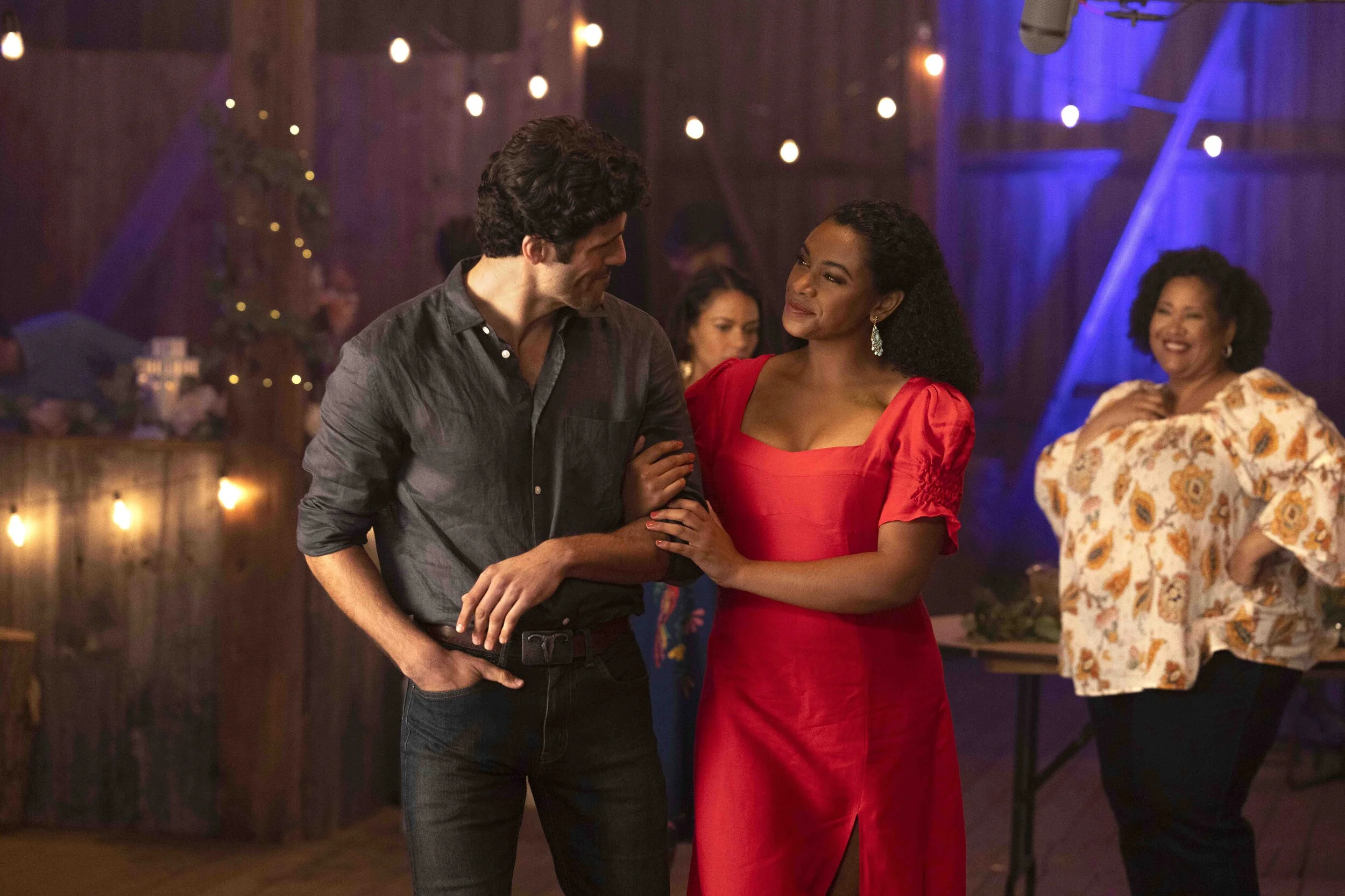 A man and woman are dancing together in a warmly lit barn, with the woman wearing a red dress and the man in a dark shirt. In the background, a woman in a floral blouse is smiling, and the barn is decorated with string lights and flowers for a celebr
