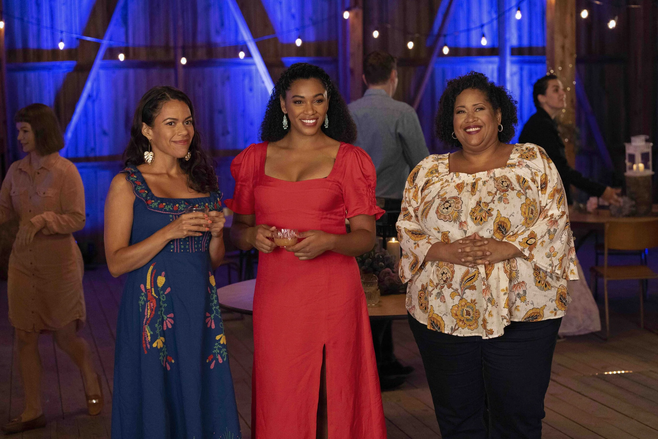 Three women standing together at a party, smiling and dressed in colorful outfits, with a rustic wooden and blue-lit background.