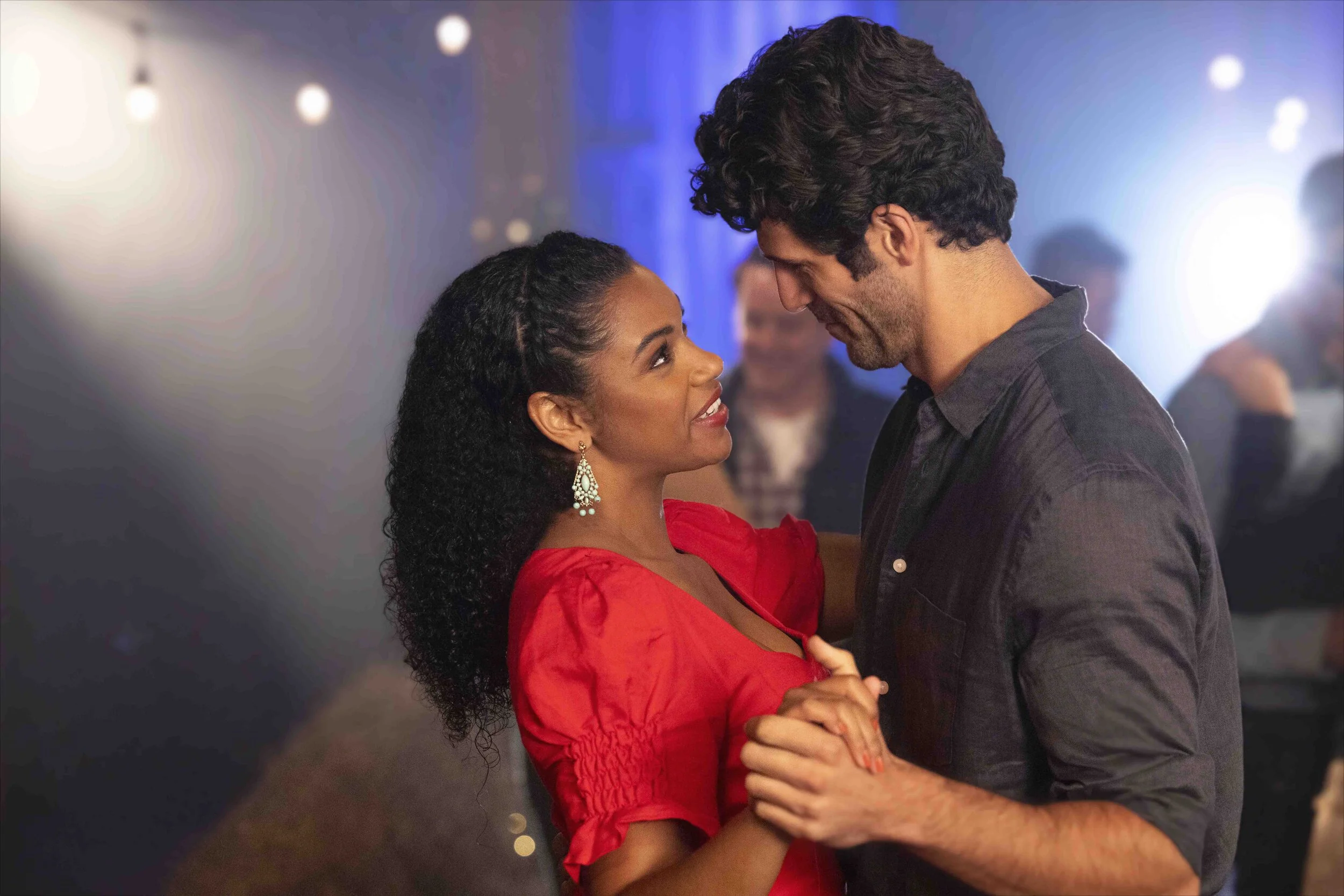 A man and woman dance closely together at a party or nightclub, looking into each other's eyes, with blurred people and colorful lights in the background.
