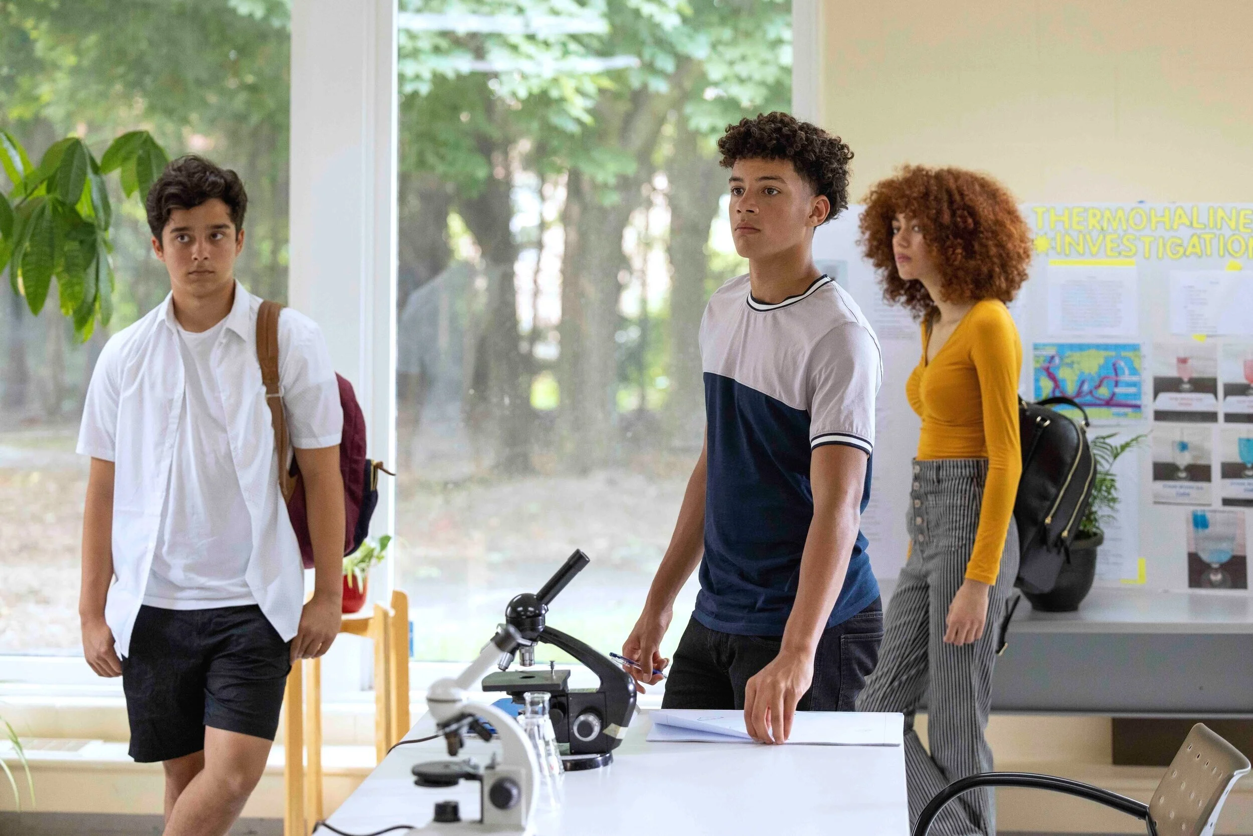 Three students in a classroom, two boys and one girl, with telescopes on a table and educational posters on the wall, during a science activity.